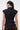 A woman with long dark hair in a ponytail wears the LILY DENIM BUTTON DOWN, a dark-wash cap-sleeve vest with a belted back, paired with brown pants with large back pockets. She faces away, highlighting the form-fitting silhouette.