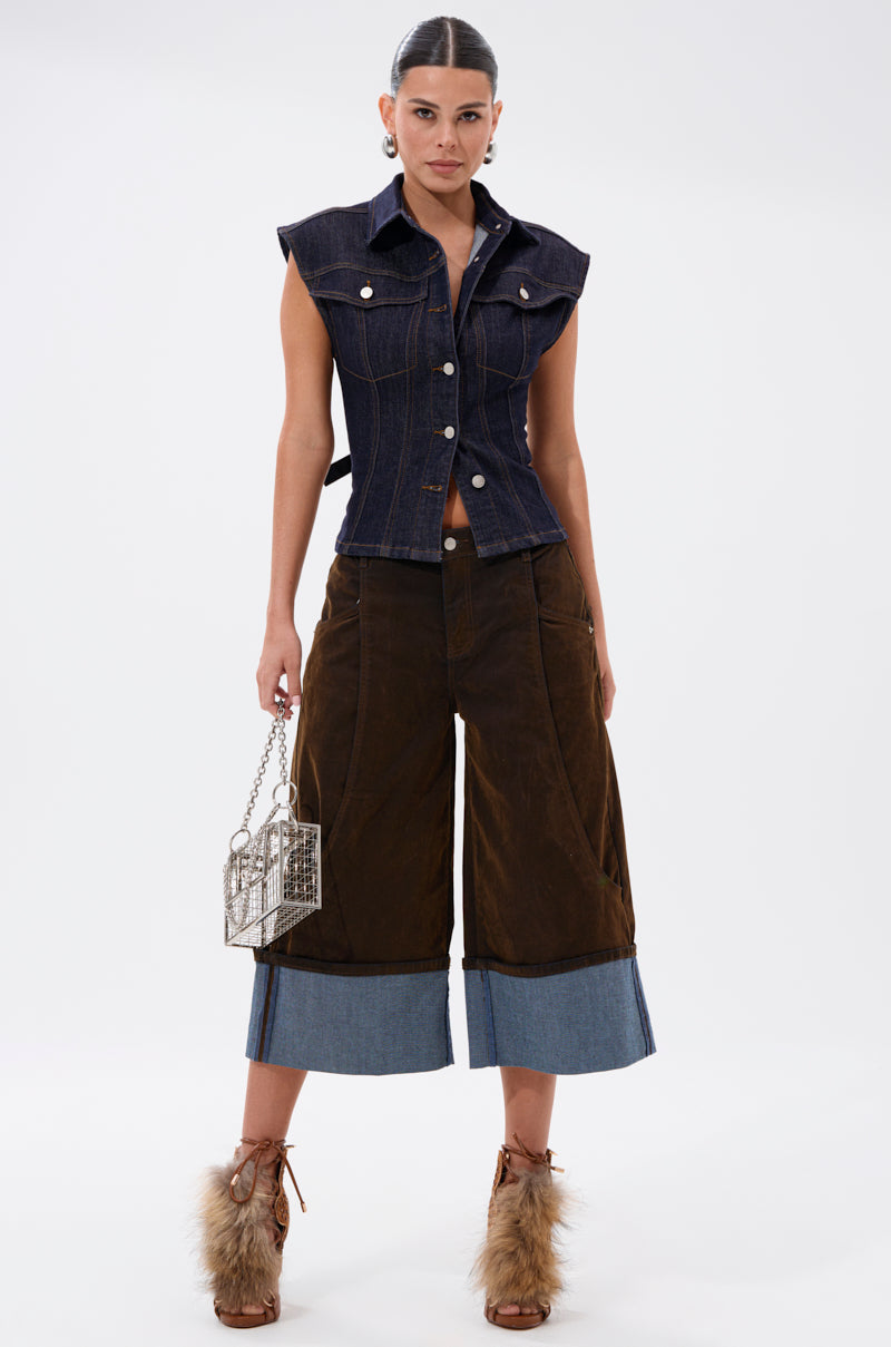 A woman poses against a white background in the LILY DENIM BUTTON DOWN, brown culotte pants with denim cuffs, furry brown ankle boots, and a metallic handbag. 8