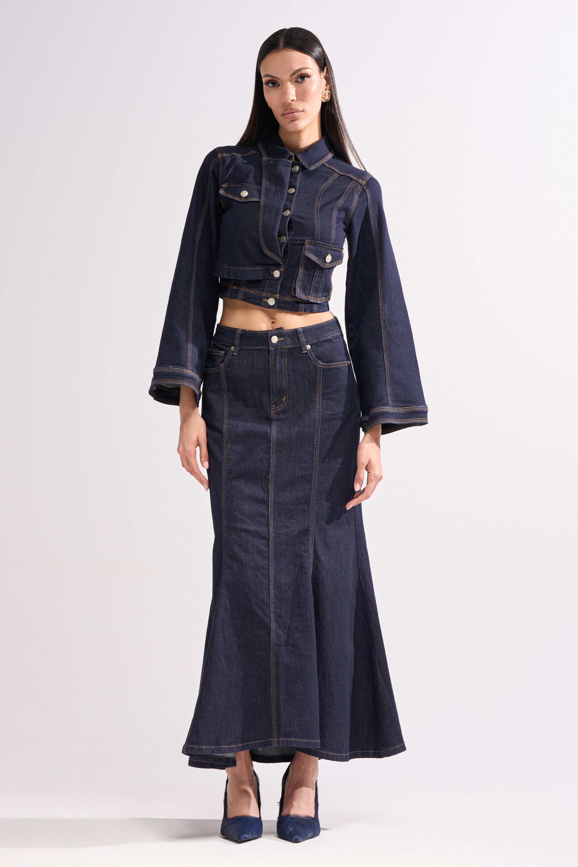 A woman wears the SHES GONE, DANCE ON DENIM MAXI SKIRT with a matching cropped long-sleeve jacket, blue high heels, and long straight hair, posing against a plain light background.