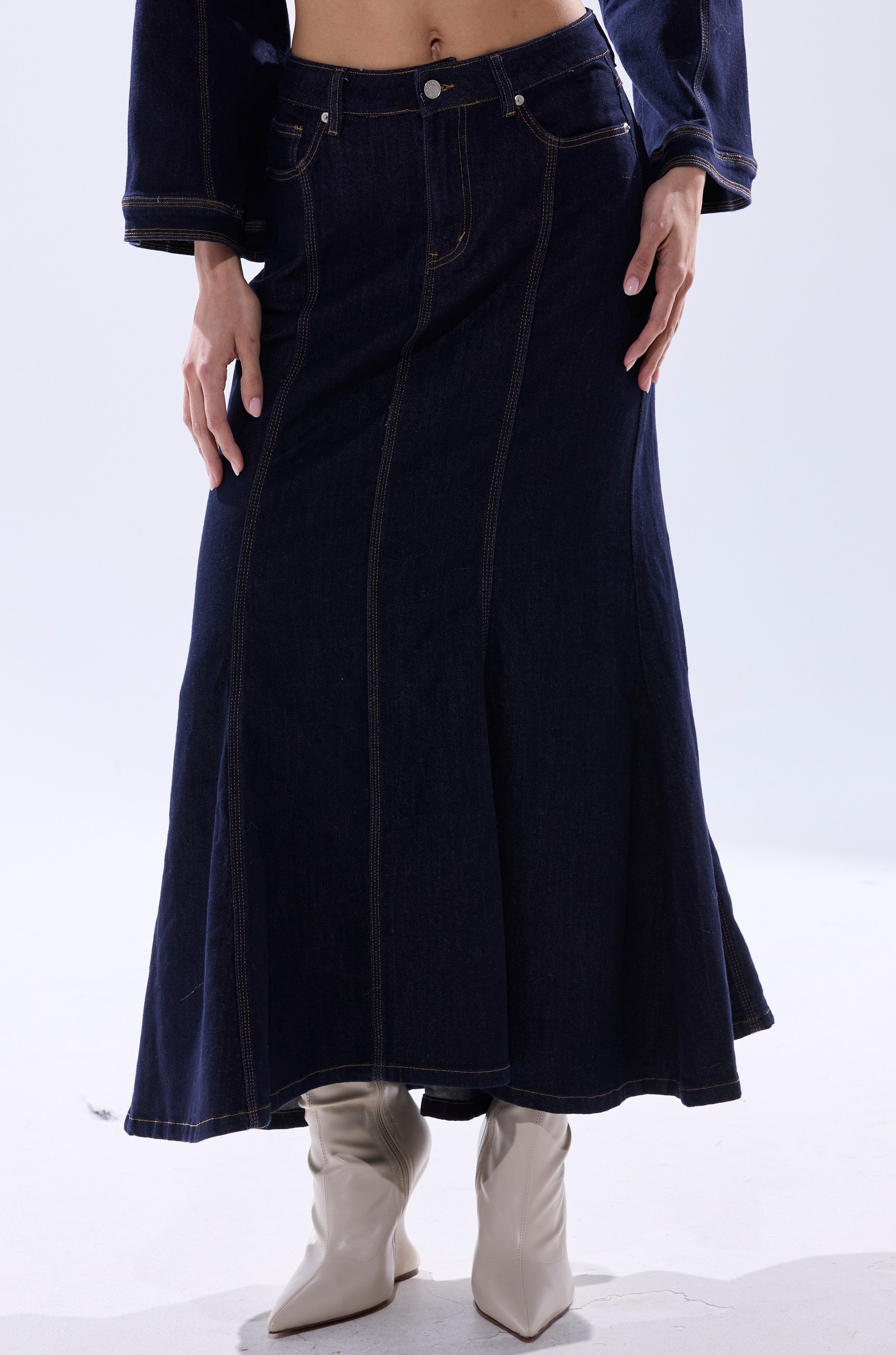 A person wears the SHES GONE, DANCE ON DENIM MAXI SKIRT with white heeled boots, their hands resting on the skirt. Only their lower body and part of their upper body are visible.
