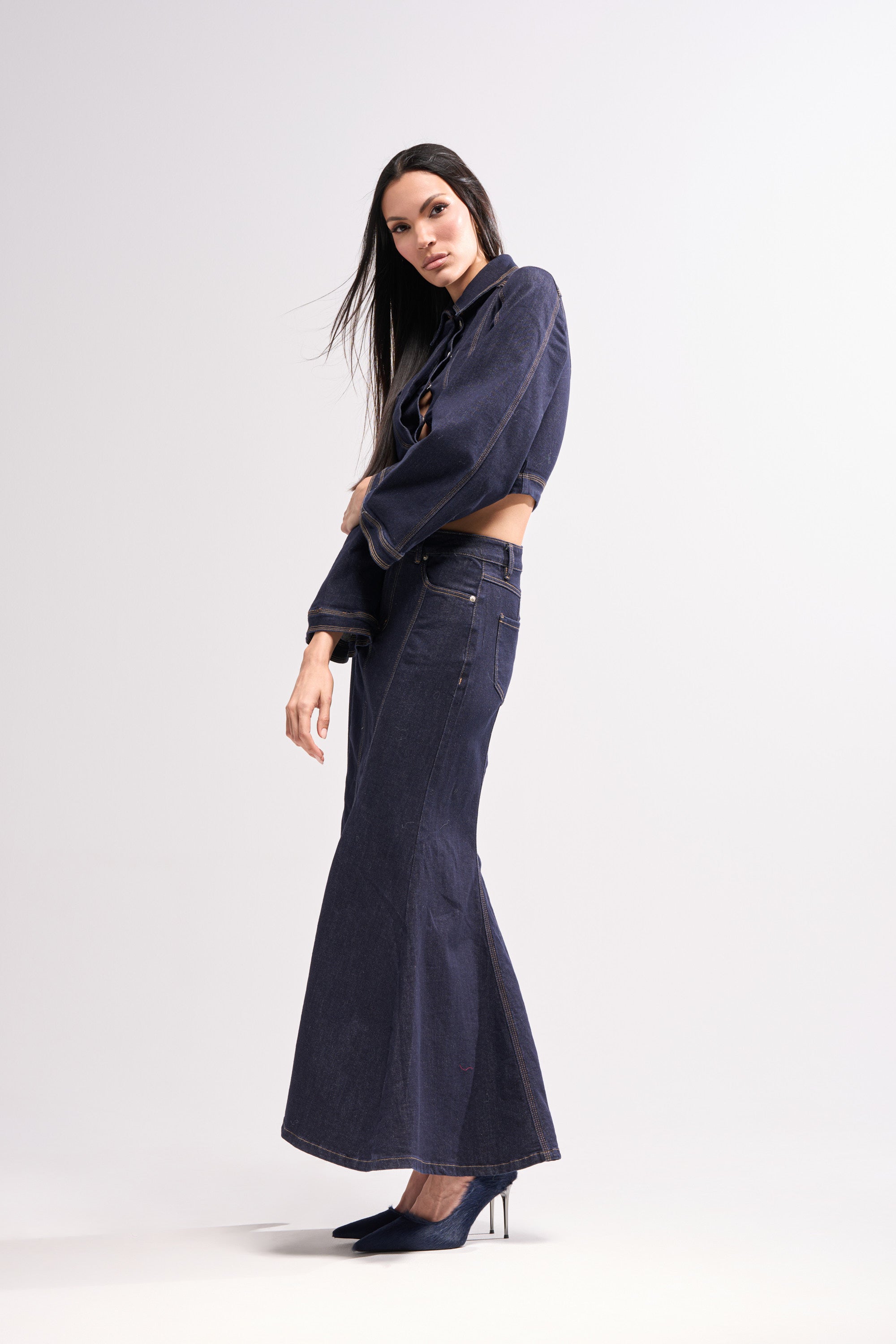 A woman poses confidently in high-heeled shoes against a plain white background, wearing the SHES GONE, DANCE ON DENIM MAXI SKIRT paired with a dark cropped denim jacket.