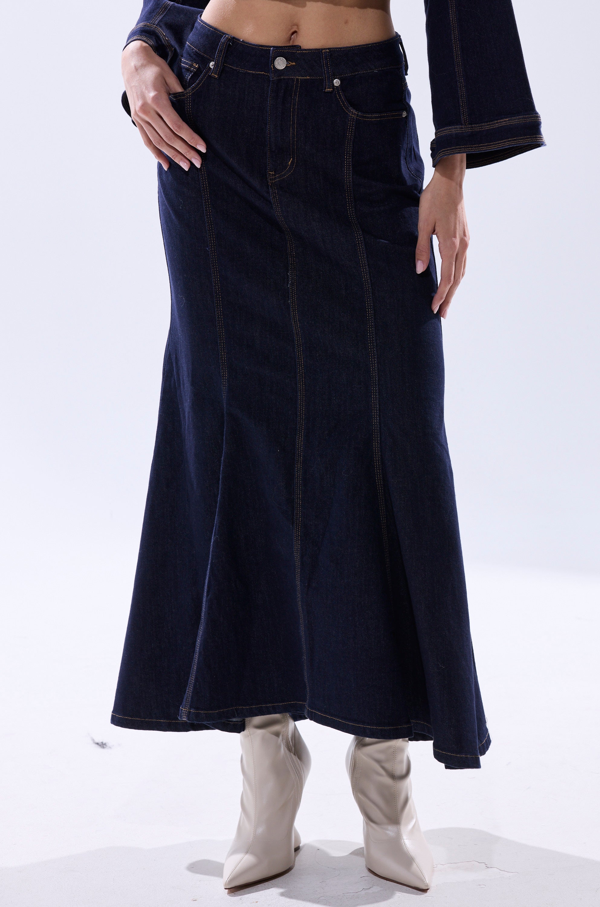 Wearing the SHES GONE, DANCE ON DENIM MAXI SKIRT in dark blue and cream pointed-toe boots, a person stands against a plain light background, one hand on their hip and the other at their side.