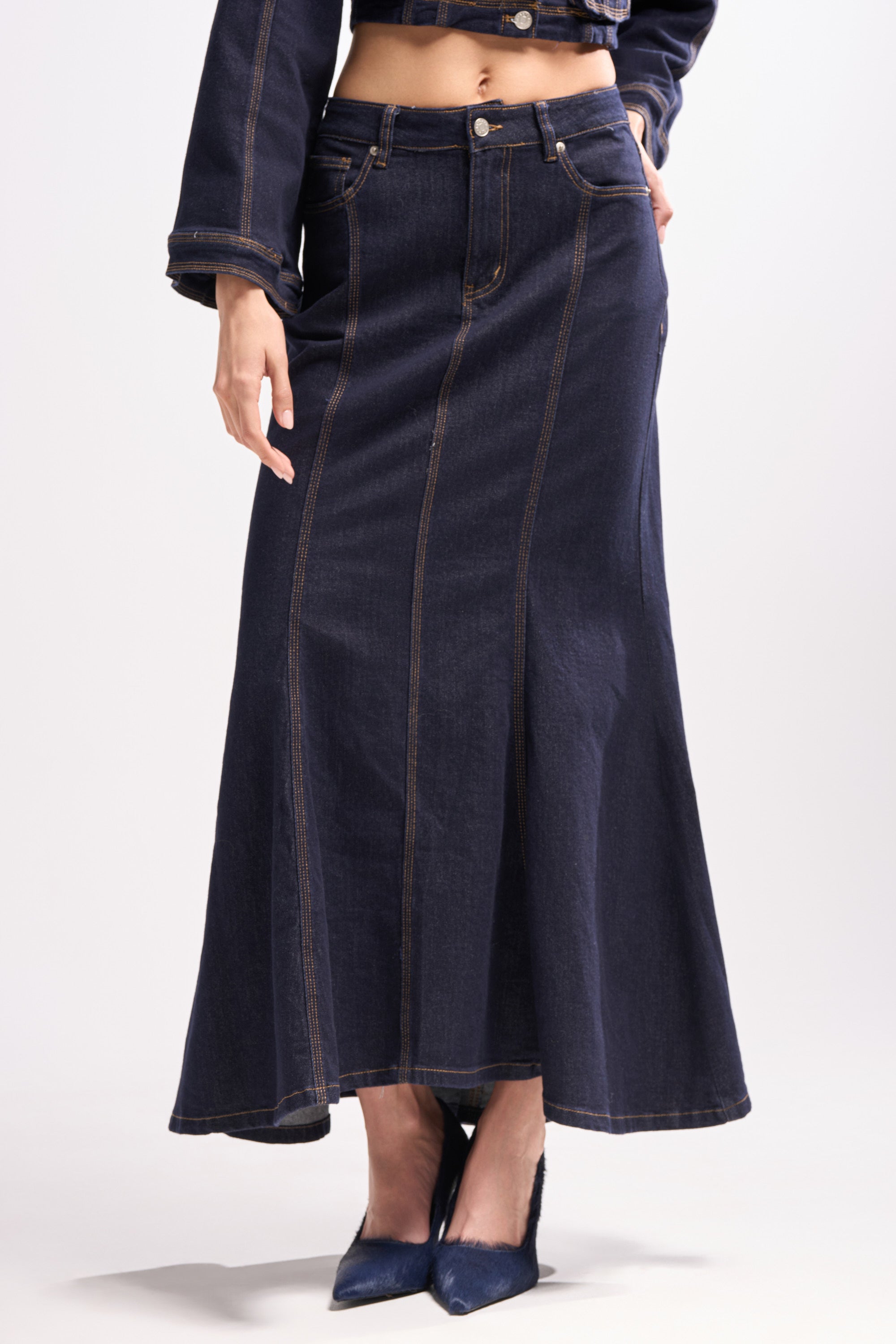 Someone wearing the SHES GONE, DANCE ON DENIM MAXI SKIRT, a dark high-rise denim maxi with visible brown stitching, styled with matching dark denim jacket sleeves and pointed-toe navy heels against a plain light background.