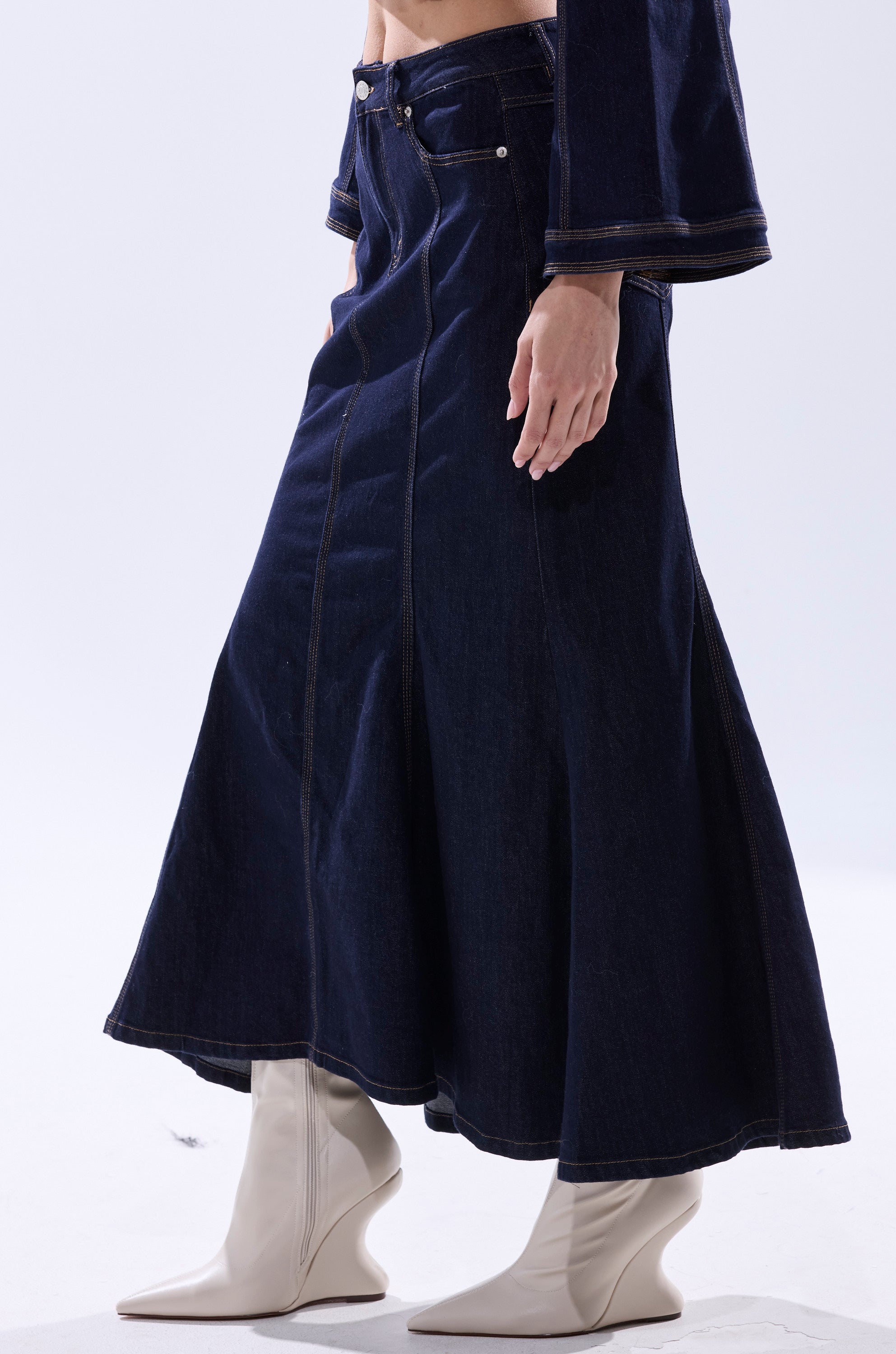 A person wears the SHES GONE, DANCE ON DENIM MAXI SKIRT—a dark high-rise denim maxi with a mermaid silhouette—paired with a matching jacket and white pointed-toe heeled boots. Flared lines and visible stitching complete the look.