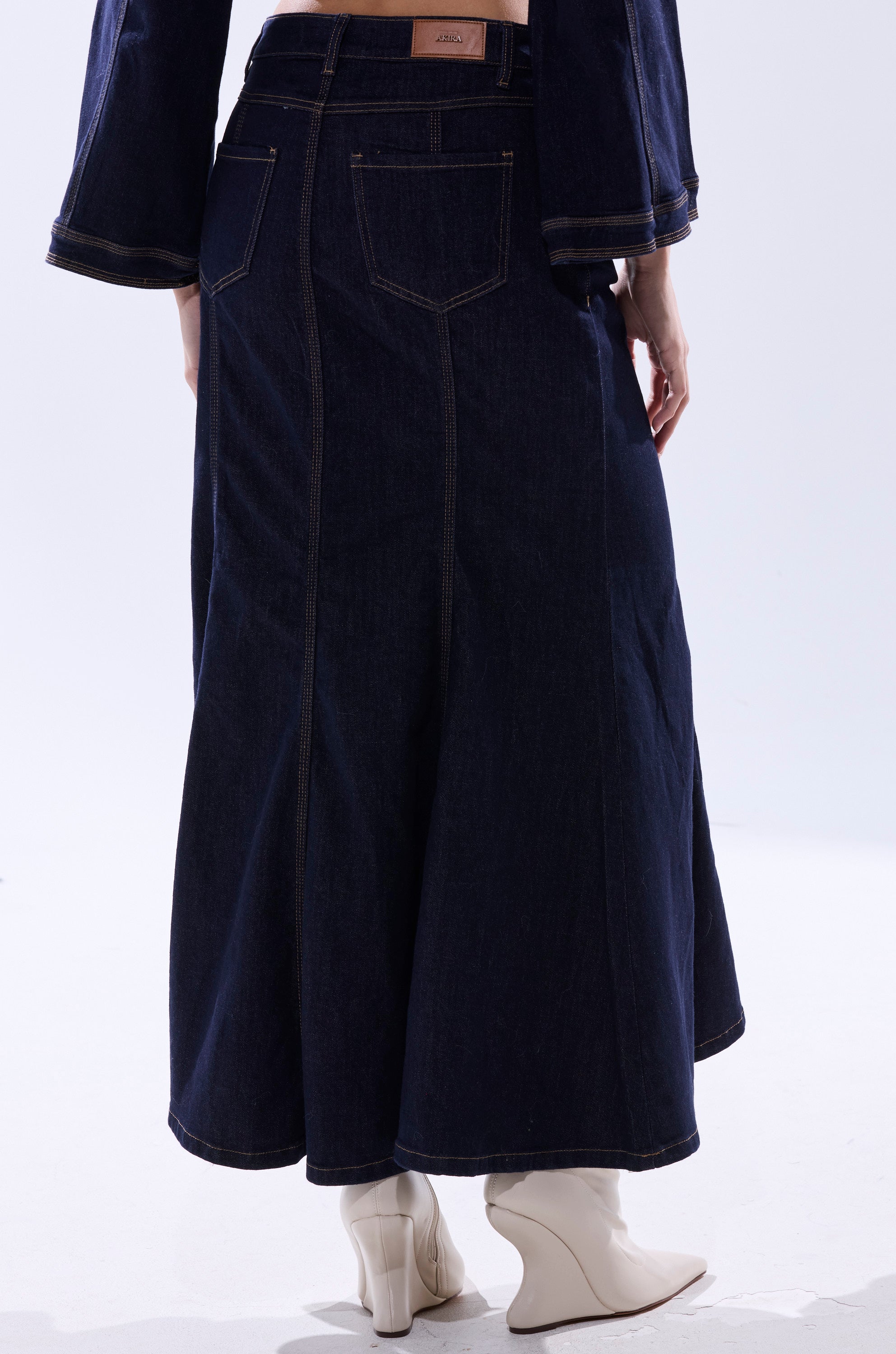 A person wearing the SHES GONE, DANCE ON DENIM MAXI SKIRT and cream heeled ankle boots stands facing away from the camera against a plain, light background.