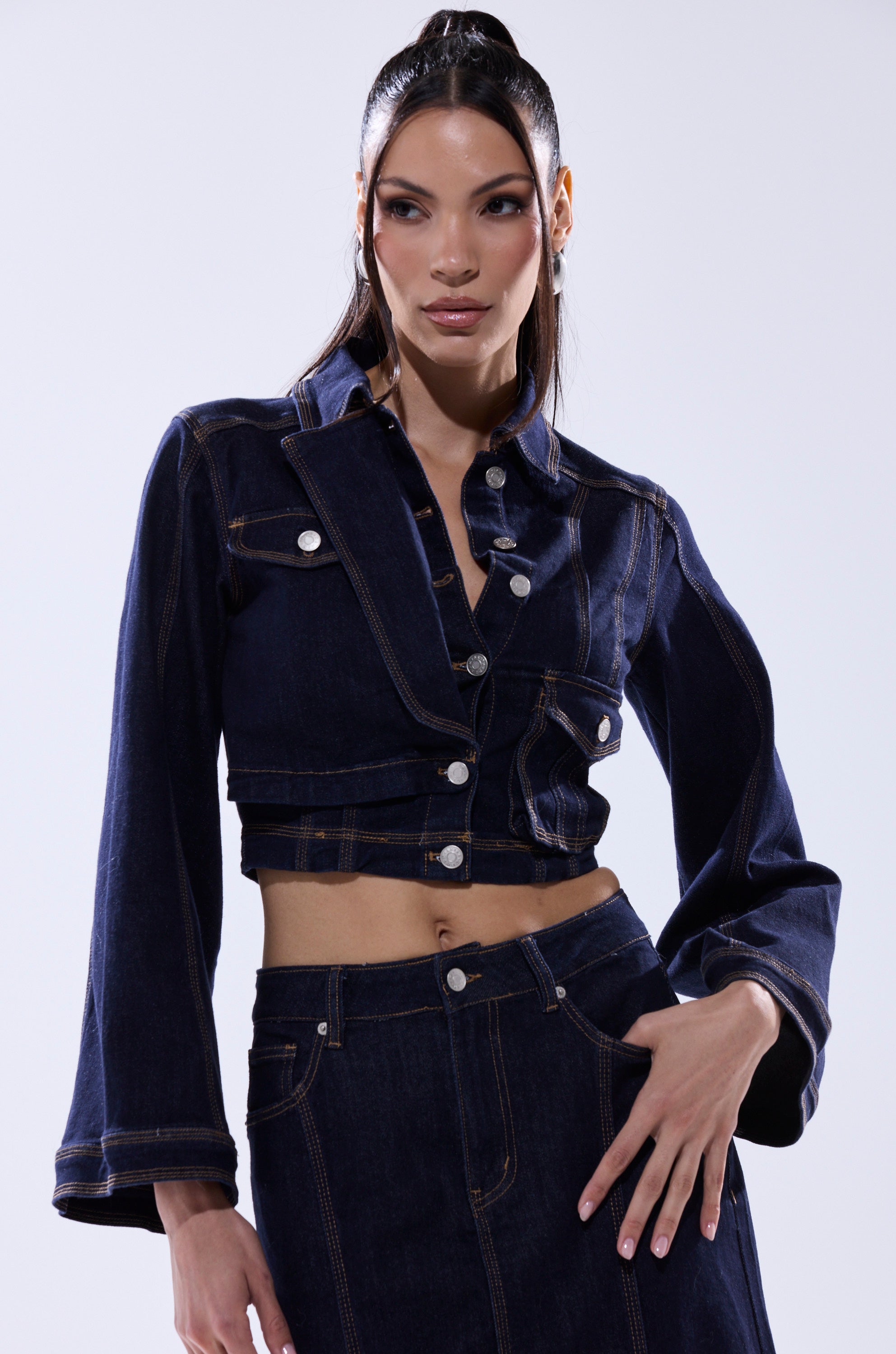 A woman poses confidently in the SHES GONE, DANCE ON DENIM BUTTON DOWN with matching high-waisted skirt against a plain light background, her long hair styled back and one hand on her hip.