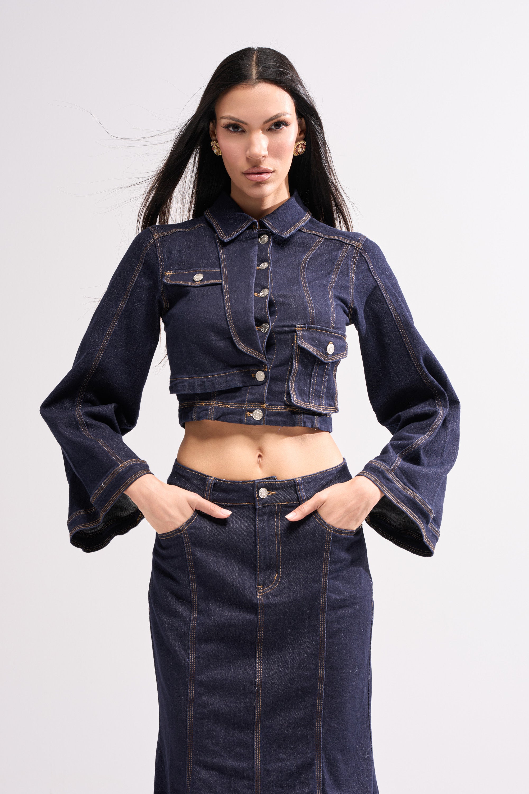 A woman with long dark hair wears the SHES GONE DANCE ON DENIM BUTTON DOWN, a dark wash cropped jacket with flared sleeves and matching high-waisted skirt, posing confidently against a plain light background.