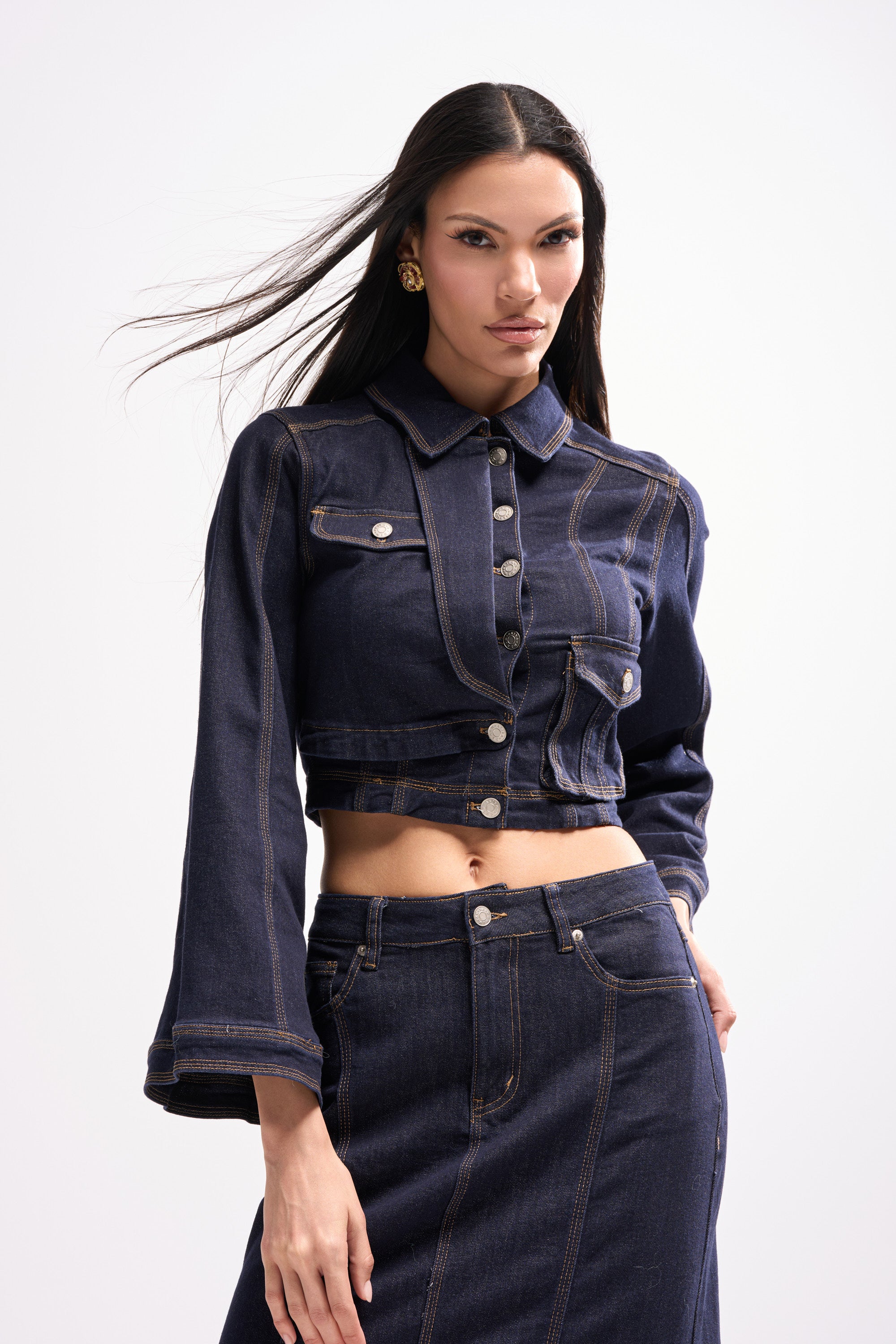A woman with long dark hair wears the SHES GONE DANCE ON DENIM BUTTON DOWN—a dark wash cropped denim jacket with silver buttons—and a matching high-waisted skirt, standing confidently against a plain light background.