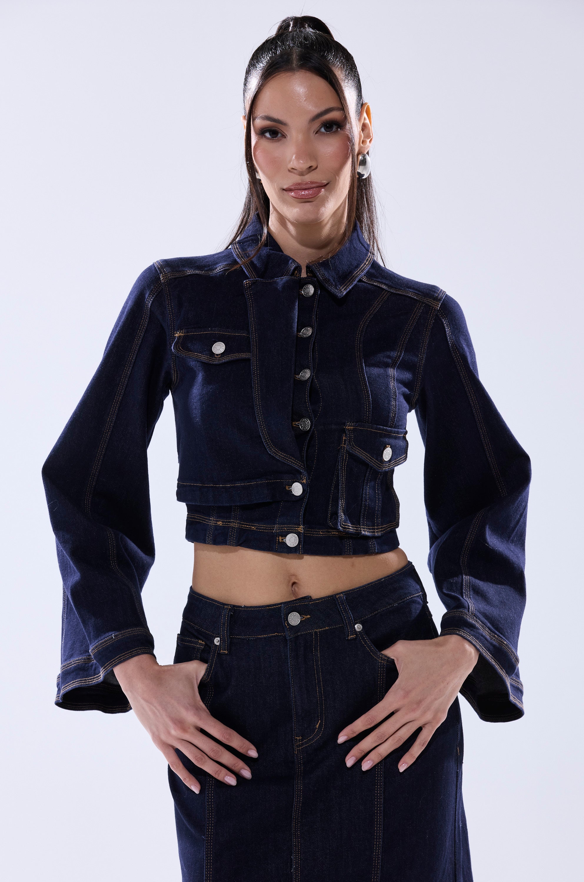 A woman confidently poses in the SHES GONE, DANCE ON DENIM BUTTON DOWN, a dark wash cropped jacket with flared sleeves, and a matching high-waisted skirt against a light background.