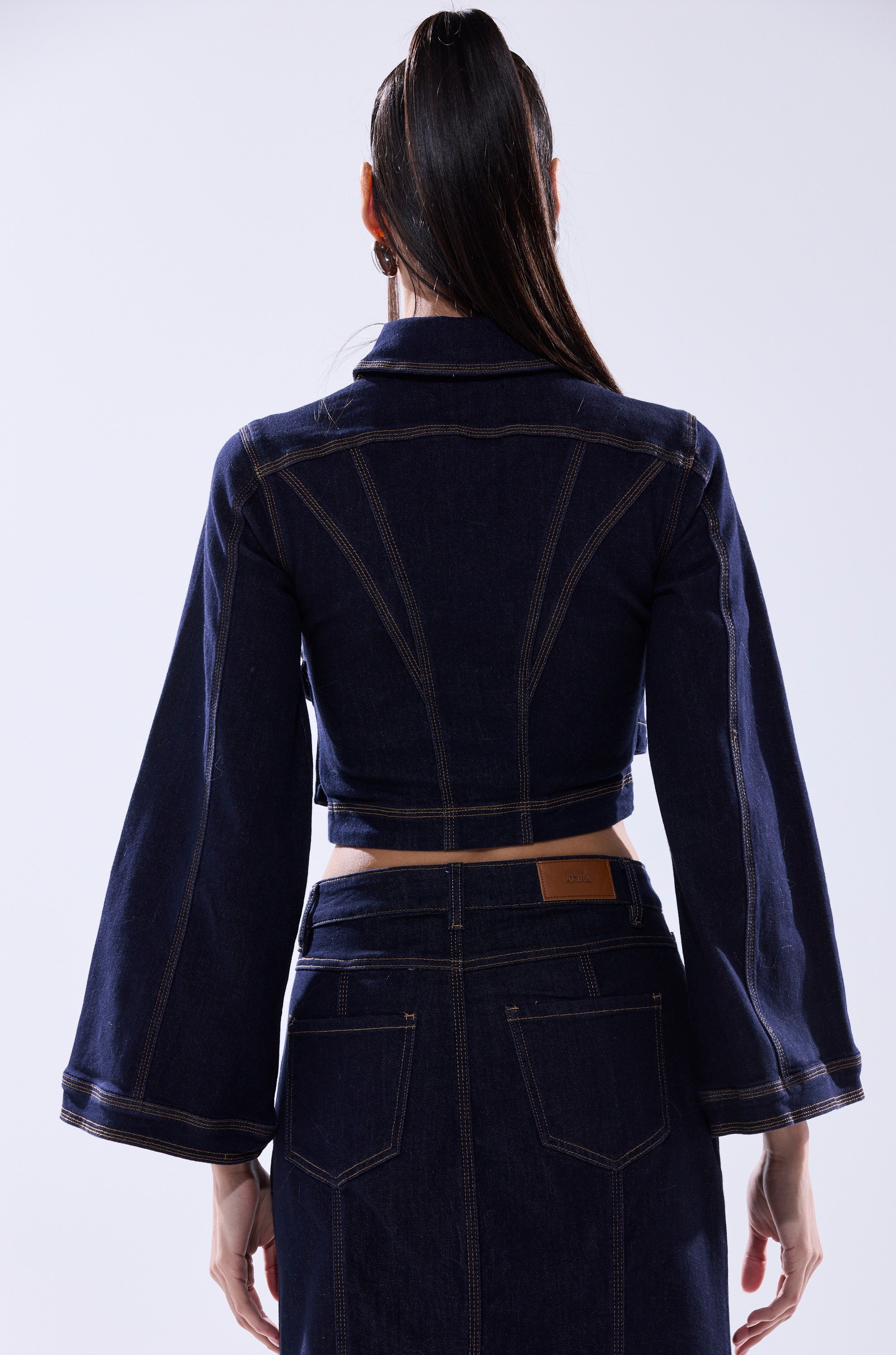 A woman with long dark hair in a ponytail is seen from behind wearing the SHES GONE DANCE ON DENIM BUTTON DOWN, a dark wash denim jacket with flared sleeves and a matching high-waisted skirt, set against a plain light-colored background.