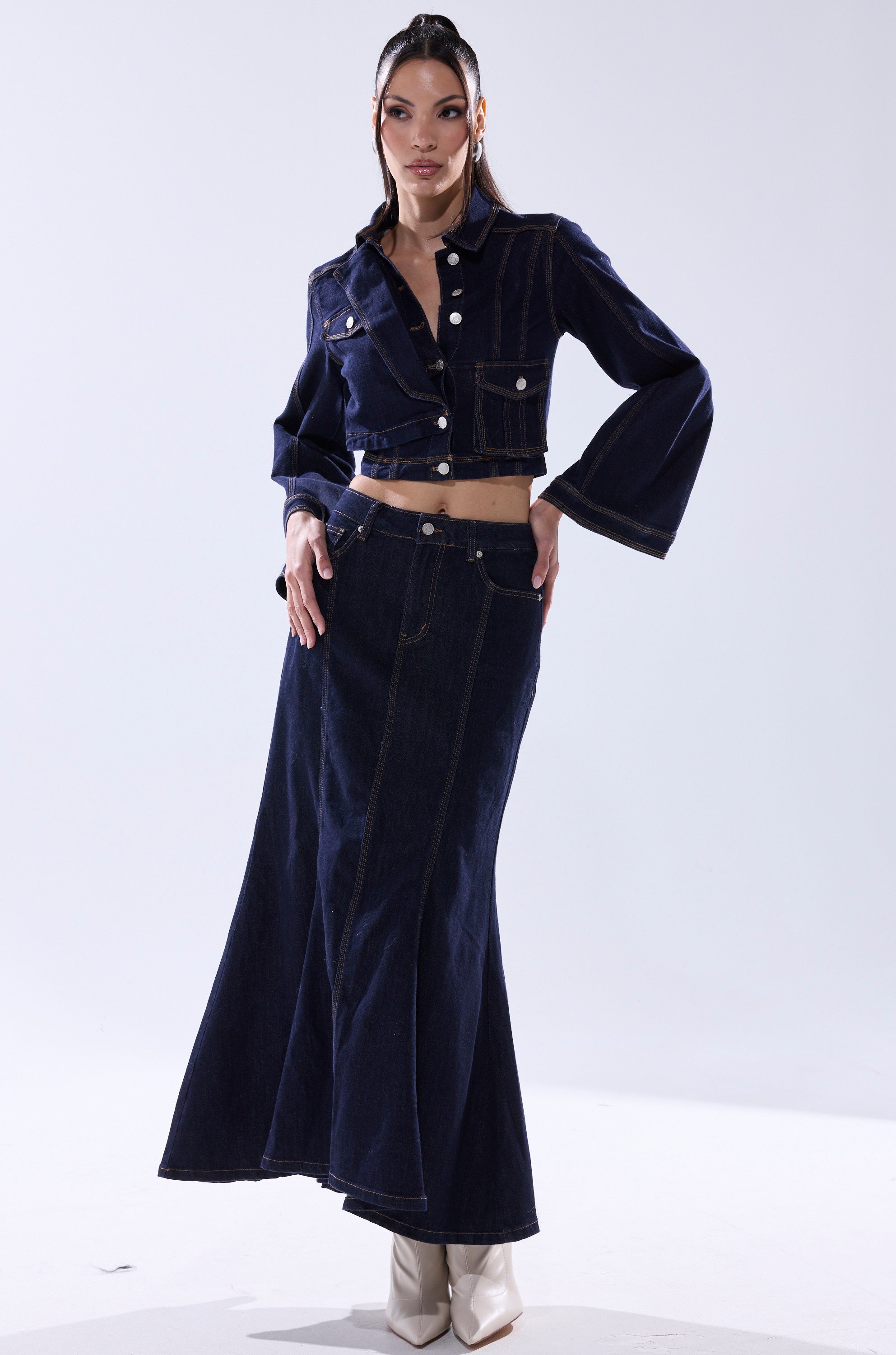 A woman wears the SHES GONE, DANCE ON DENIM BUTTON DOWN— a cropped, wide-sleeve dark wash denim jacket styled with a high-waisted flared long skirt and white ankle boots.