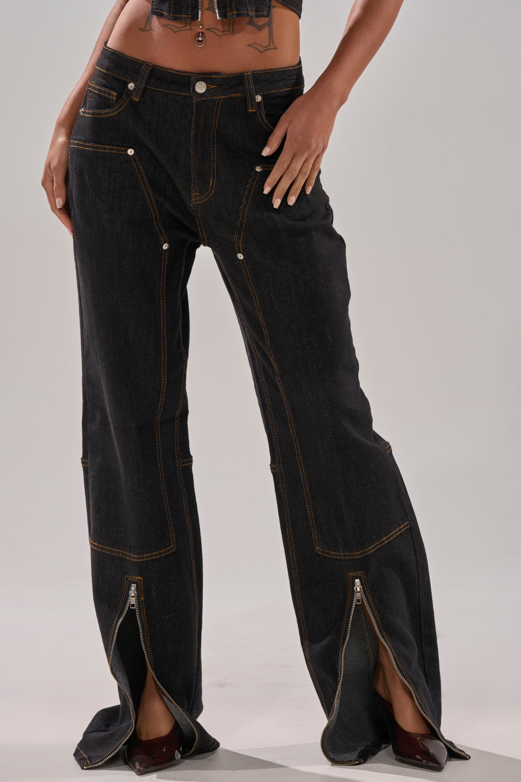 Wearing the HARDEST TO LOVE WIDE LEG DENIM, high-rise dark-wash jeans with visible stitching and front zippers partly unzipped at the hem to show pointed brown shoes, this person stands with hands on hips.
