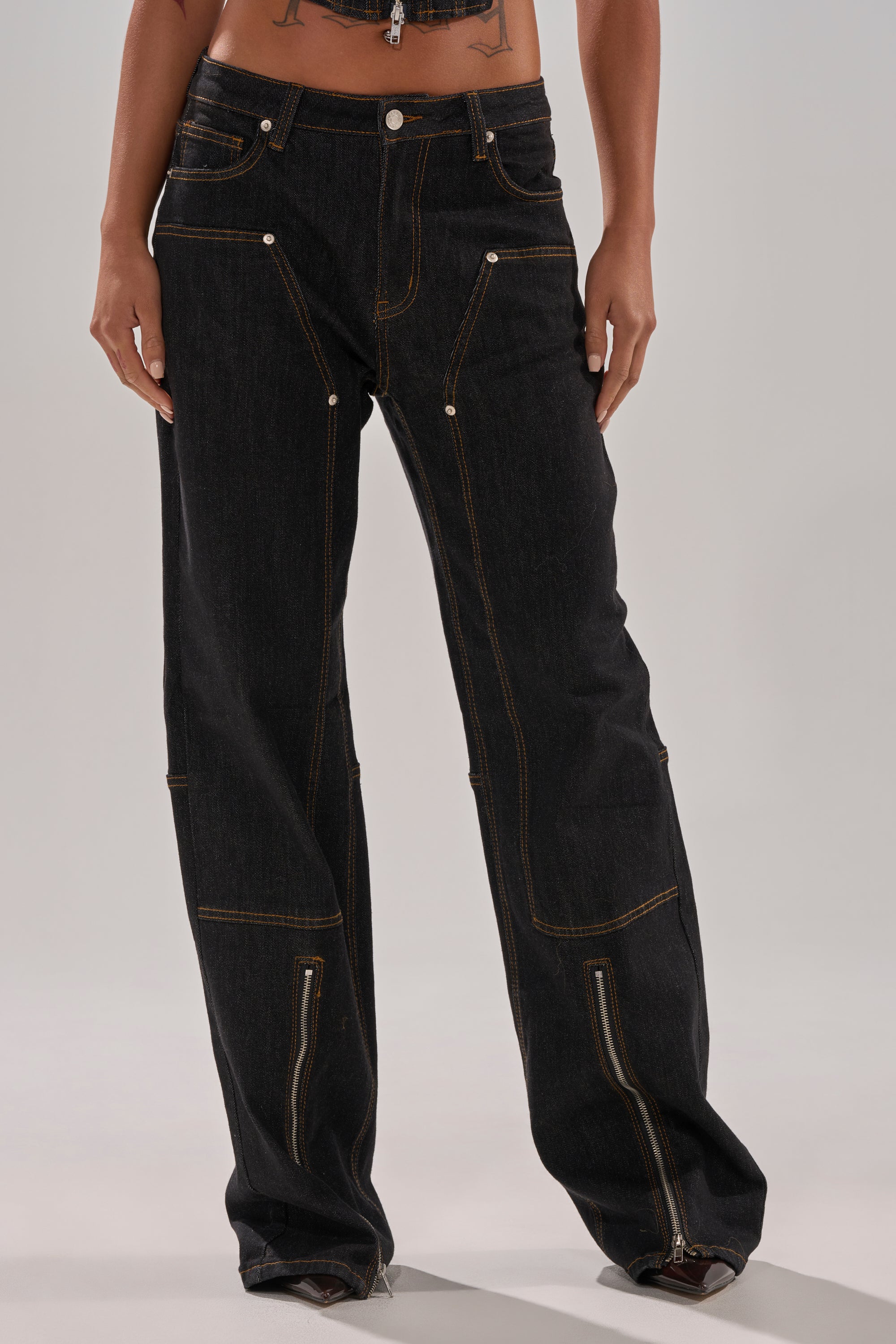 A person wears HARDEST TO LOVE WIDE LEG DENIM—loose, high-rise dark-wash jeans featuring gold stitching, silver buttons, and exposed ankle zippers—standing against a plain light background.