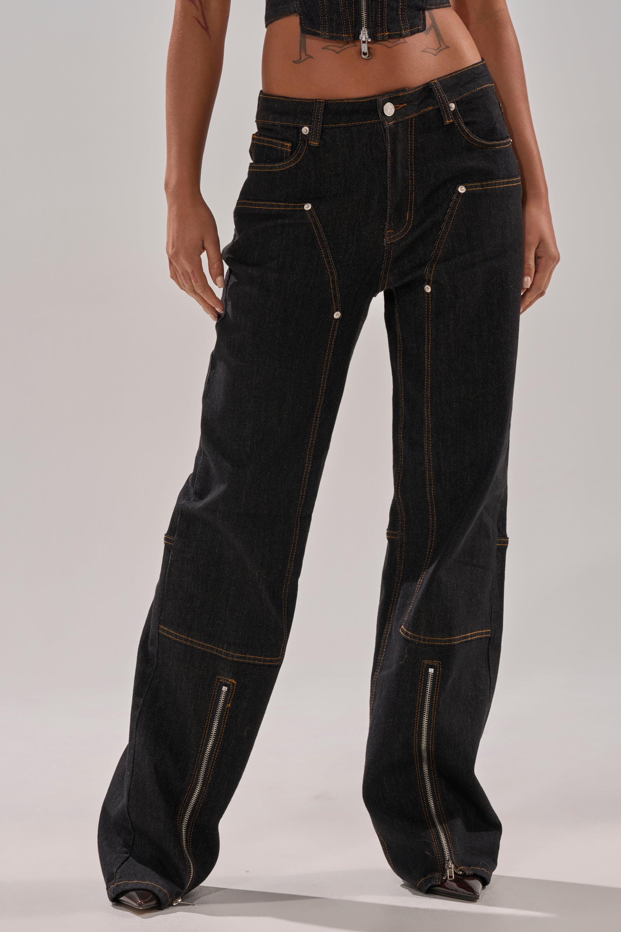 Wearing HARDEST TO LOVE WIDE LEG DENIM—high-rise, loose-fitting black jeans with brown stitching, multiple pockets, and lower leg zippers—paired with a matching cropped top. Only the lower torso and legs are shown.
