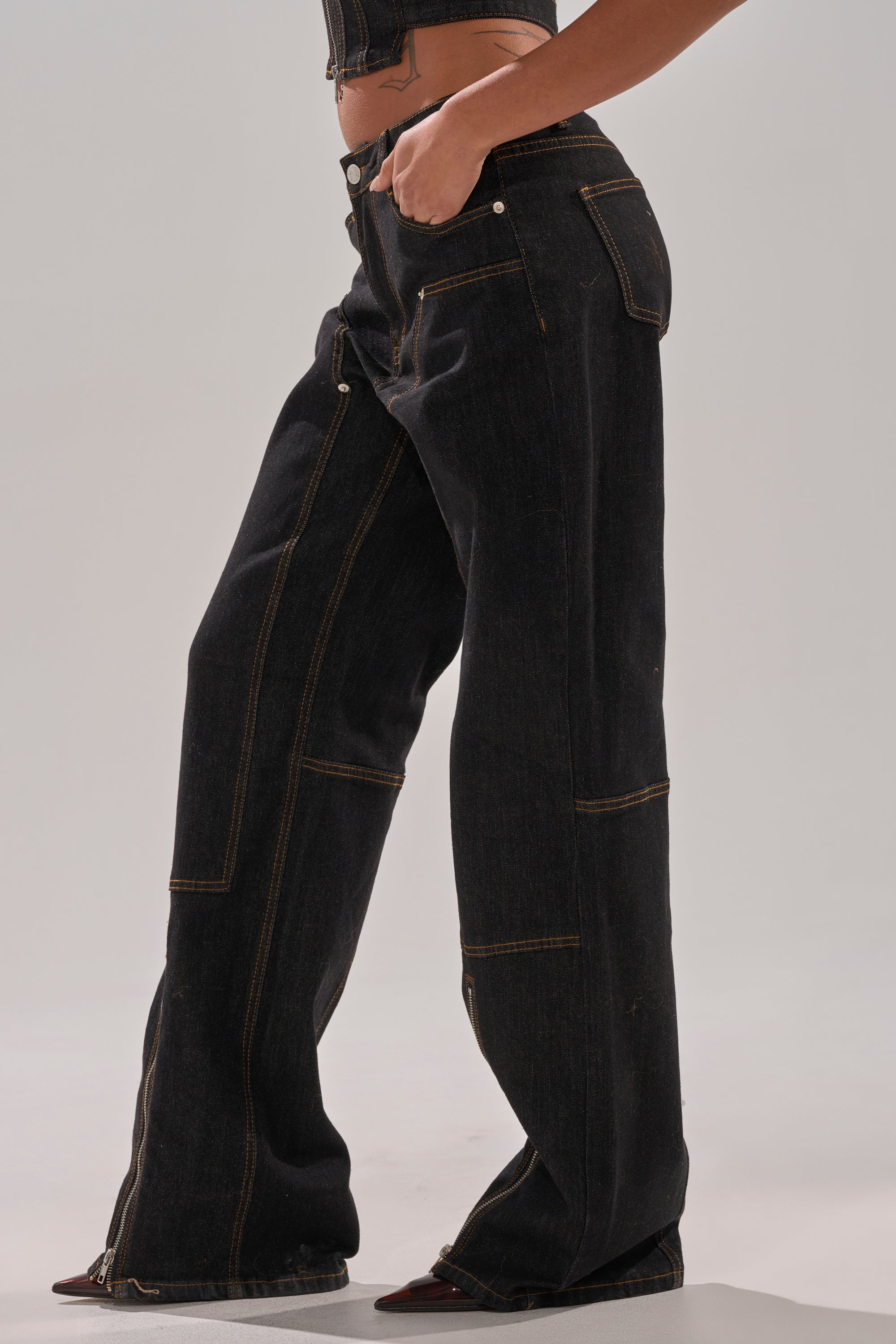 A person models the HARDEST TO LOVE WIDE LEG DENIM—high-rise, wide leg jeans in a dark wash with bold stitching and side pockets—shown from mid-torso to feet against a plain background.