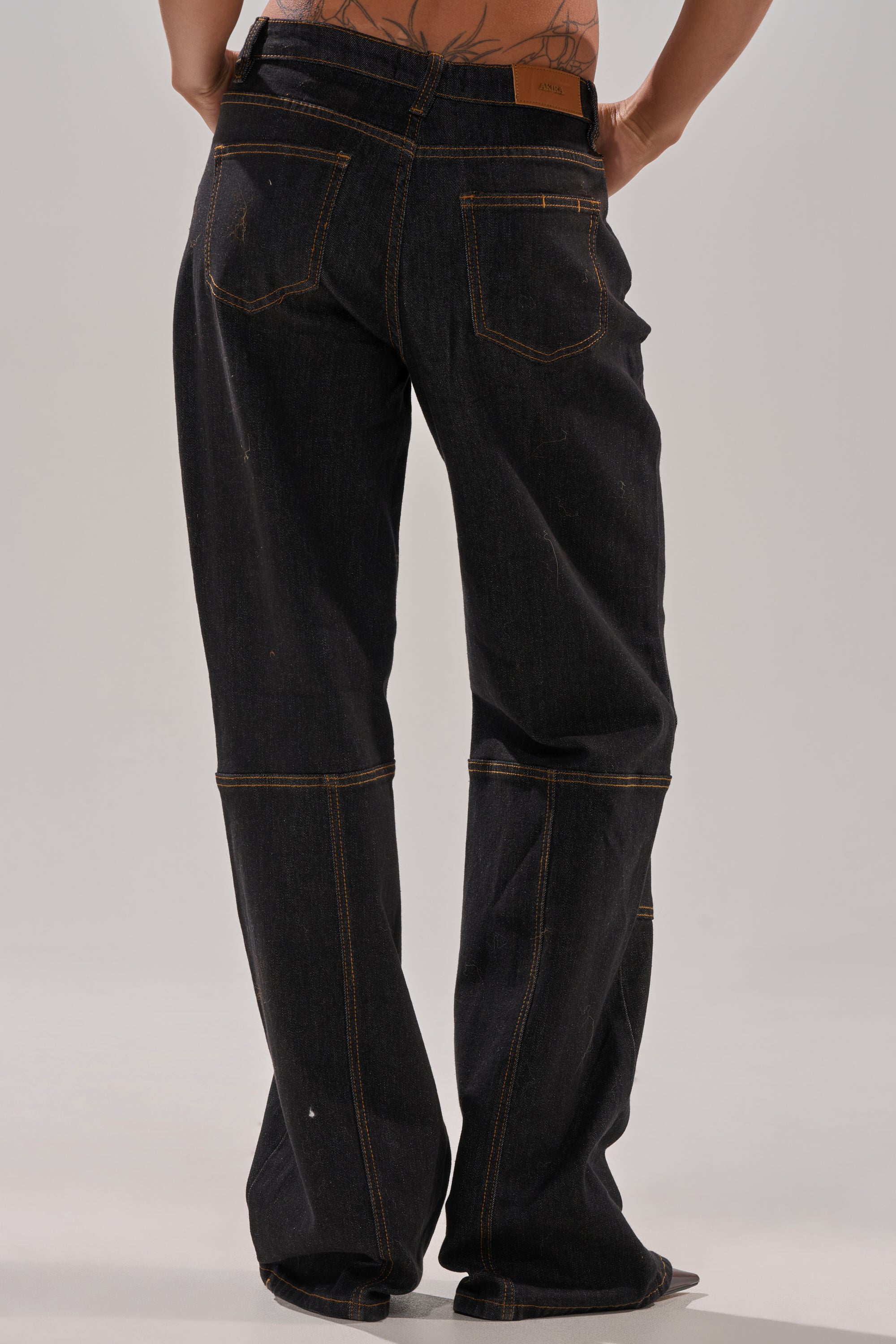 Someone stands facing away, partially showing their torso and arms, wearing HARDEST TO LOVE WIDE LEG DENIM—loose-fitting, high-rise jeans in dark wash denim with yellow stitching, emphasizing the back pockets and waistband.