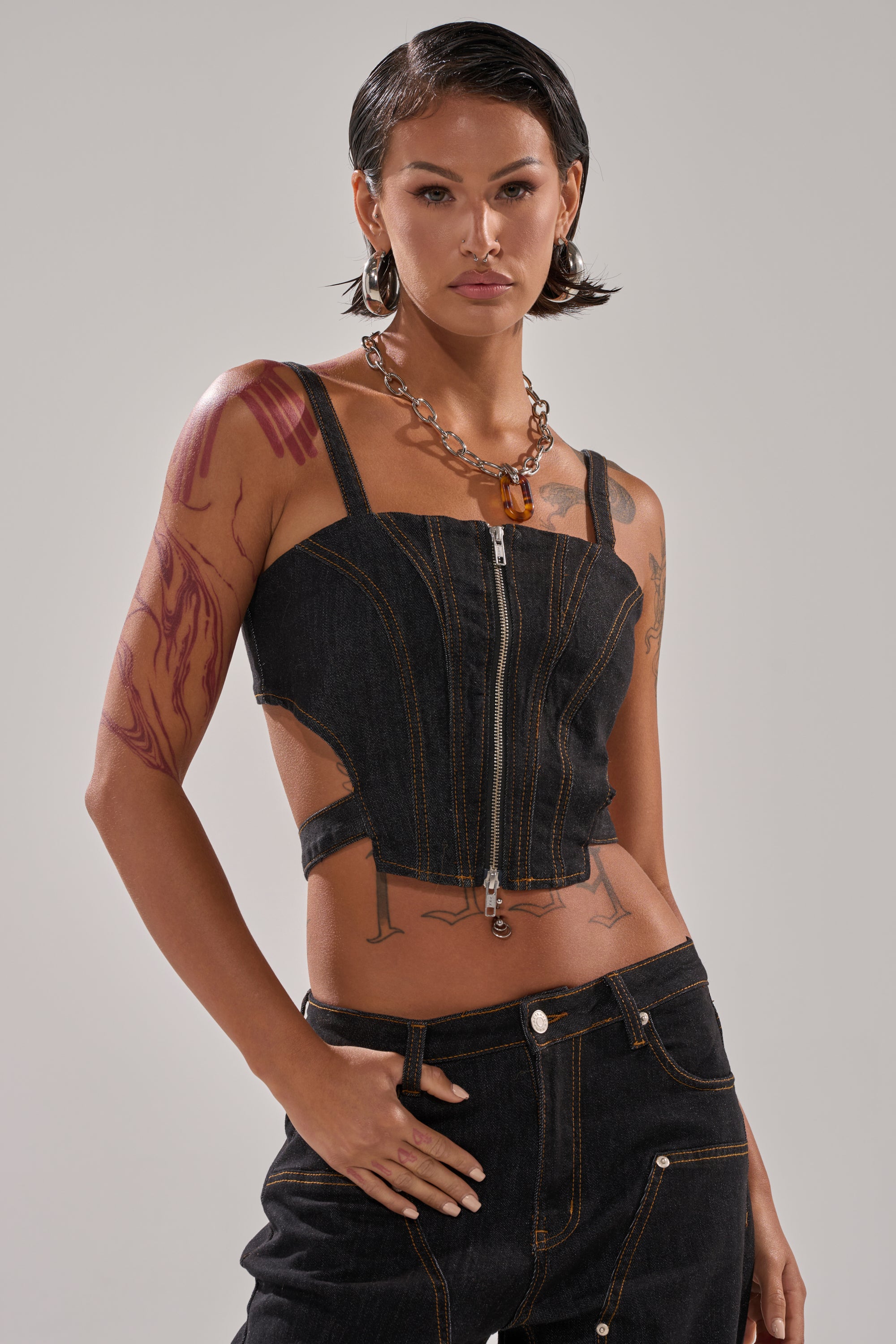 A person with short dark hair wears the HARDEST TO LOVE DENIM CORSET and matching jeans, highlighting a curve-hugging fit. They pose with one hand in their pocket, accessorized with large hoop earrings, a chunky necklace, and arm tattoos.