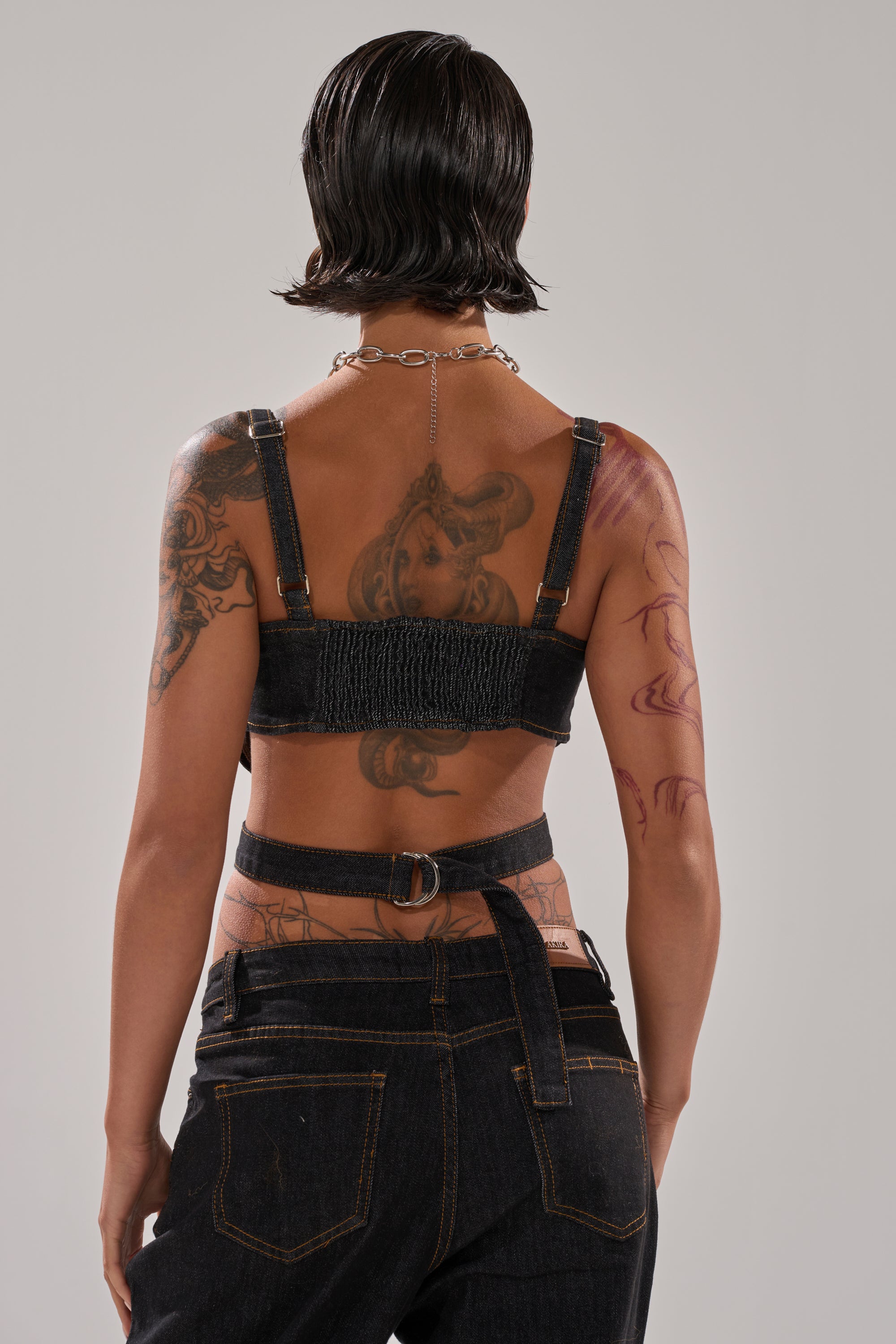 A person with short, slicked-back hair is seen from behind, wearing the HARDEST TO LOVE DENIM CORSET and matching black pants. Their tattooed back and arms are complemented by a silver chain necklace.
