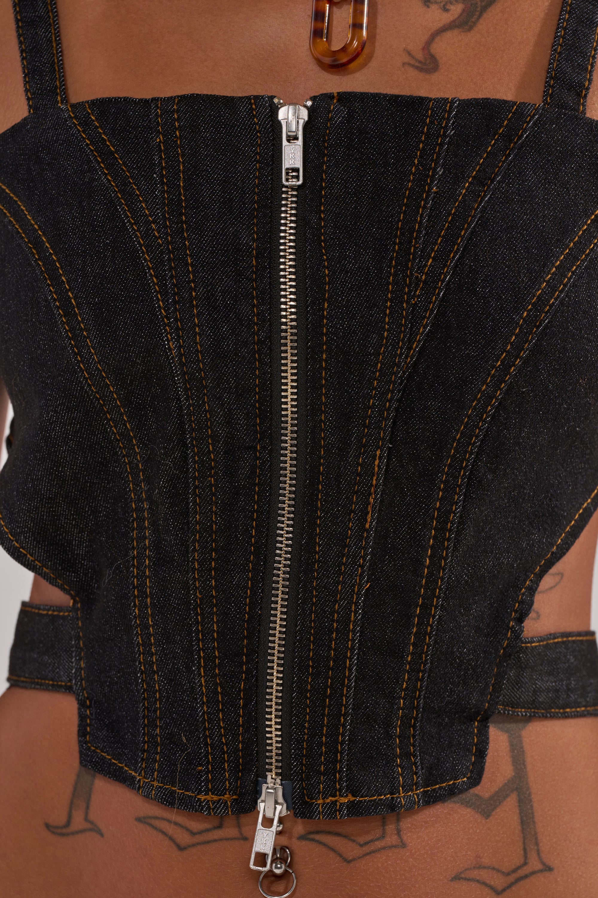 Close-up of a person wearing the HARDEST TO LOVE DENIM CORSET—black denim with orange stitching and a front zipper. Tattoos cover their chest and stomach, complemented by a brown translucent pendant necklace.
