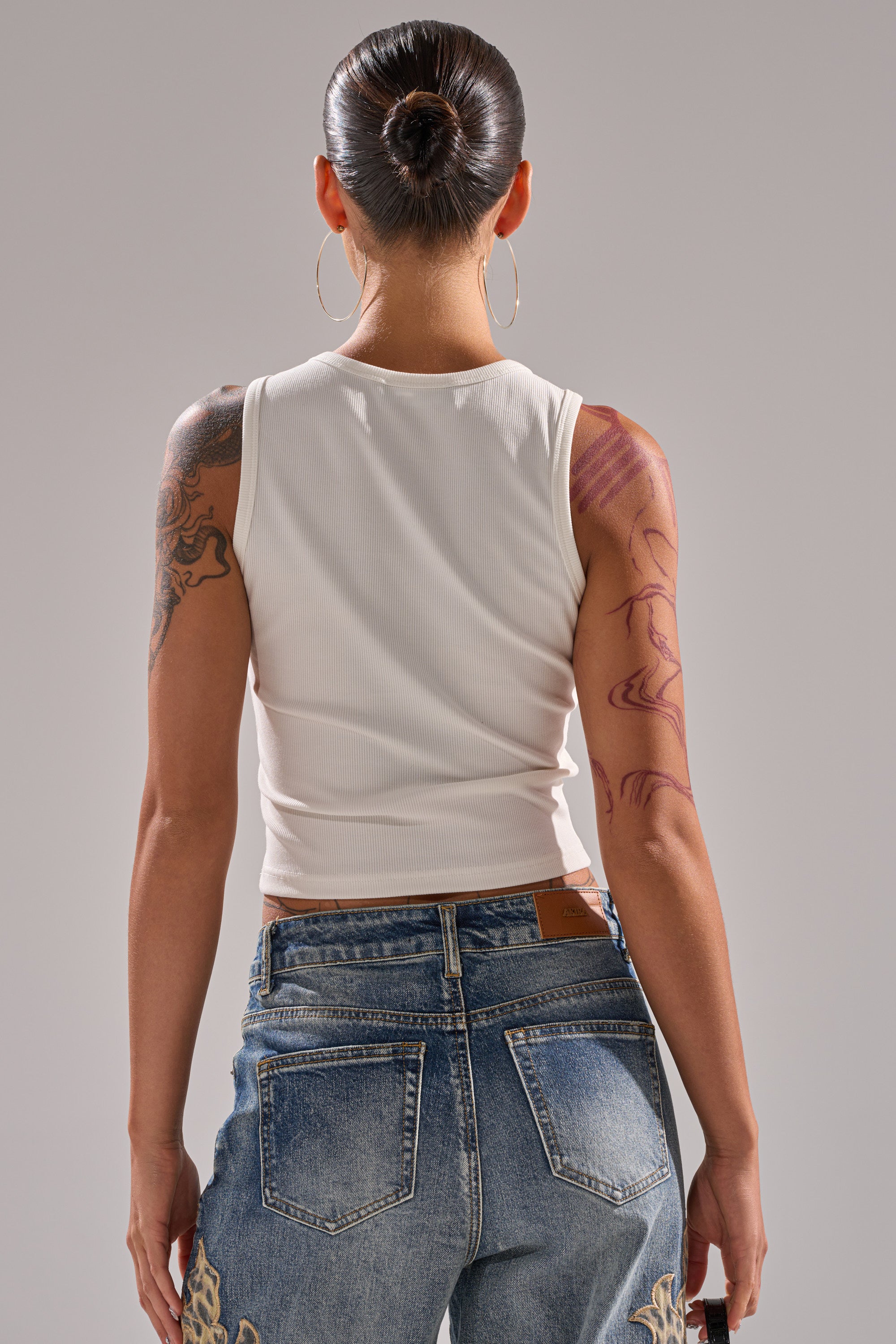 A woman with dark hair in a bun, wearing large hoop earrings and the LOSER BYE TANK with blue jeans, faces away to display her colorful arm and back tattoos, all set against a neutral gray background. 4