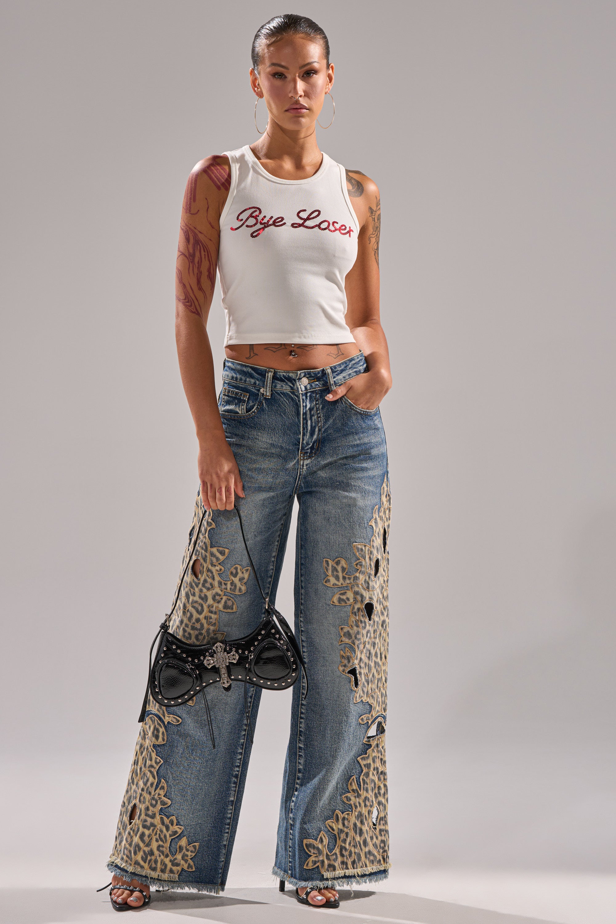 A woman stands confidently in the LOSER BYE TANK, a ribbed knit top with bold red text, paired with wide-leg blue jeans with lace cutouts, sandals, and a black handbag accented with a bow. 5