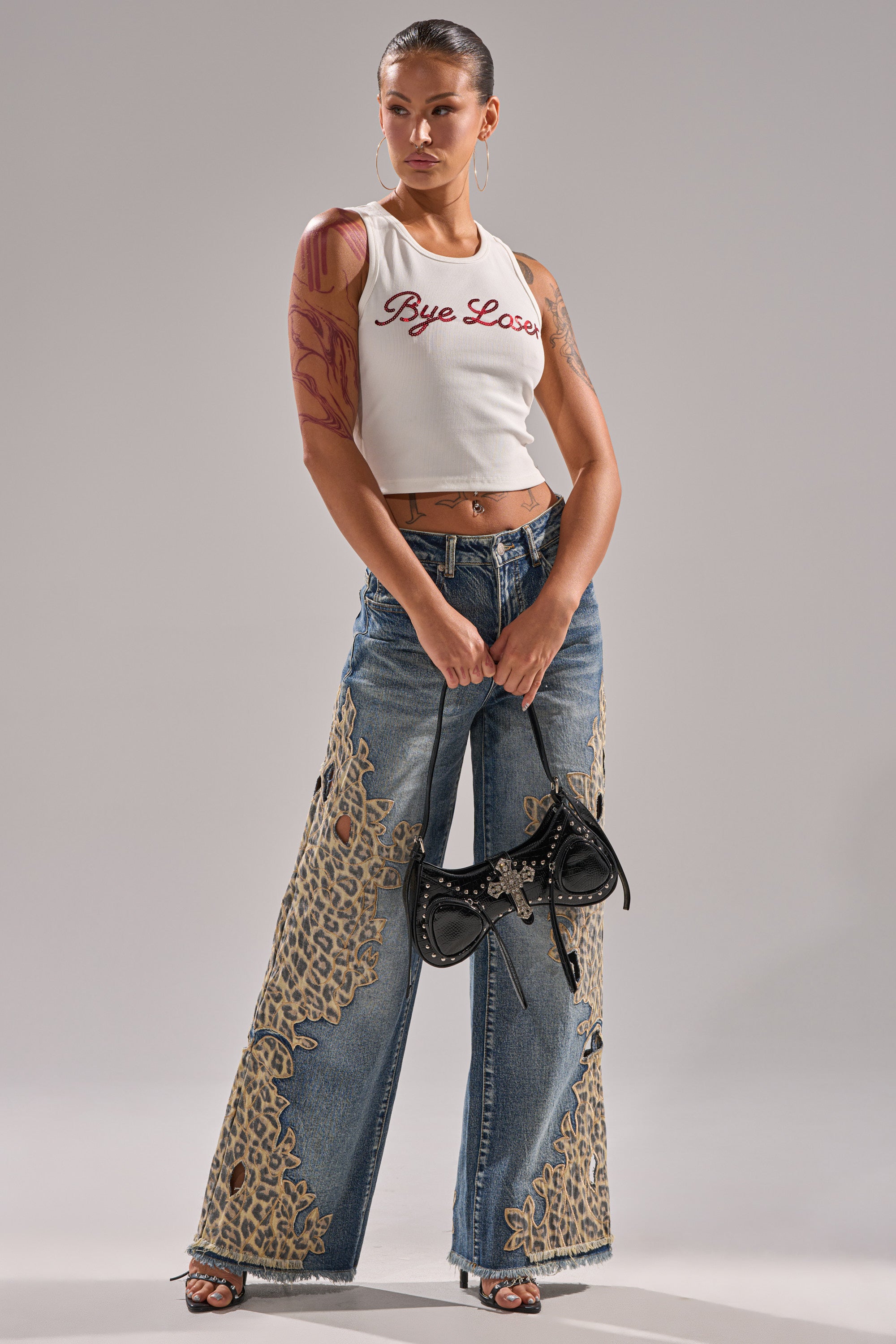 A woman stands confidently in the white, ribbed-knit LOSER BYE TANK with bold red text, styled with blue wide-leg jeans with beige lace accents, black heeled sandals, a black mini handbag, slicked-back hair, and a visible arm tattoo. 6