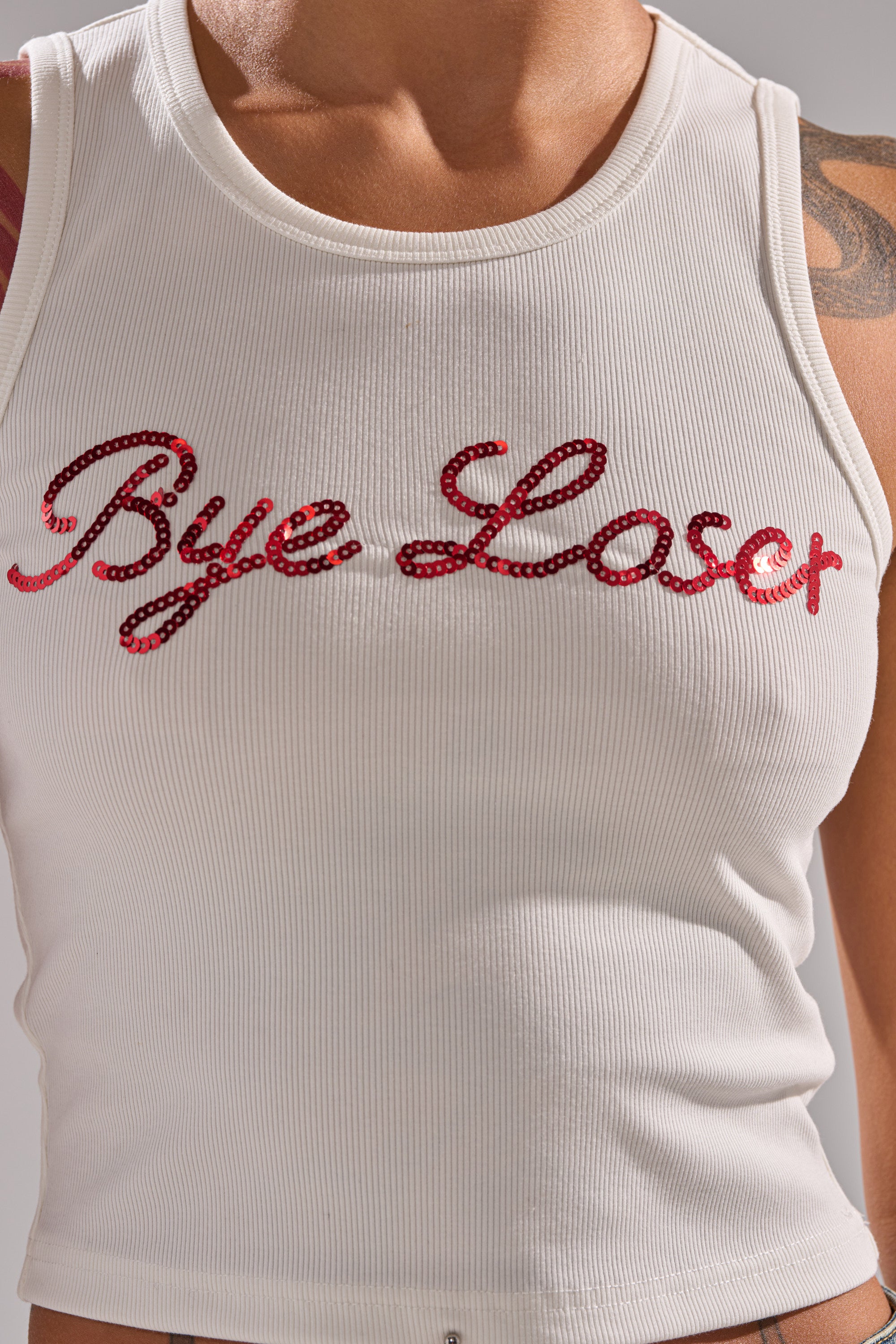 Someone wears the LOSER BYE TANK, a white ribbed knit tank top with red sequin lettering spelling "Bye Loser." The image shows only the torso and part of one arm. 7