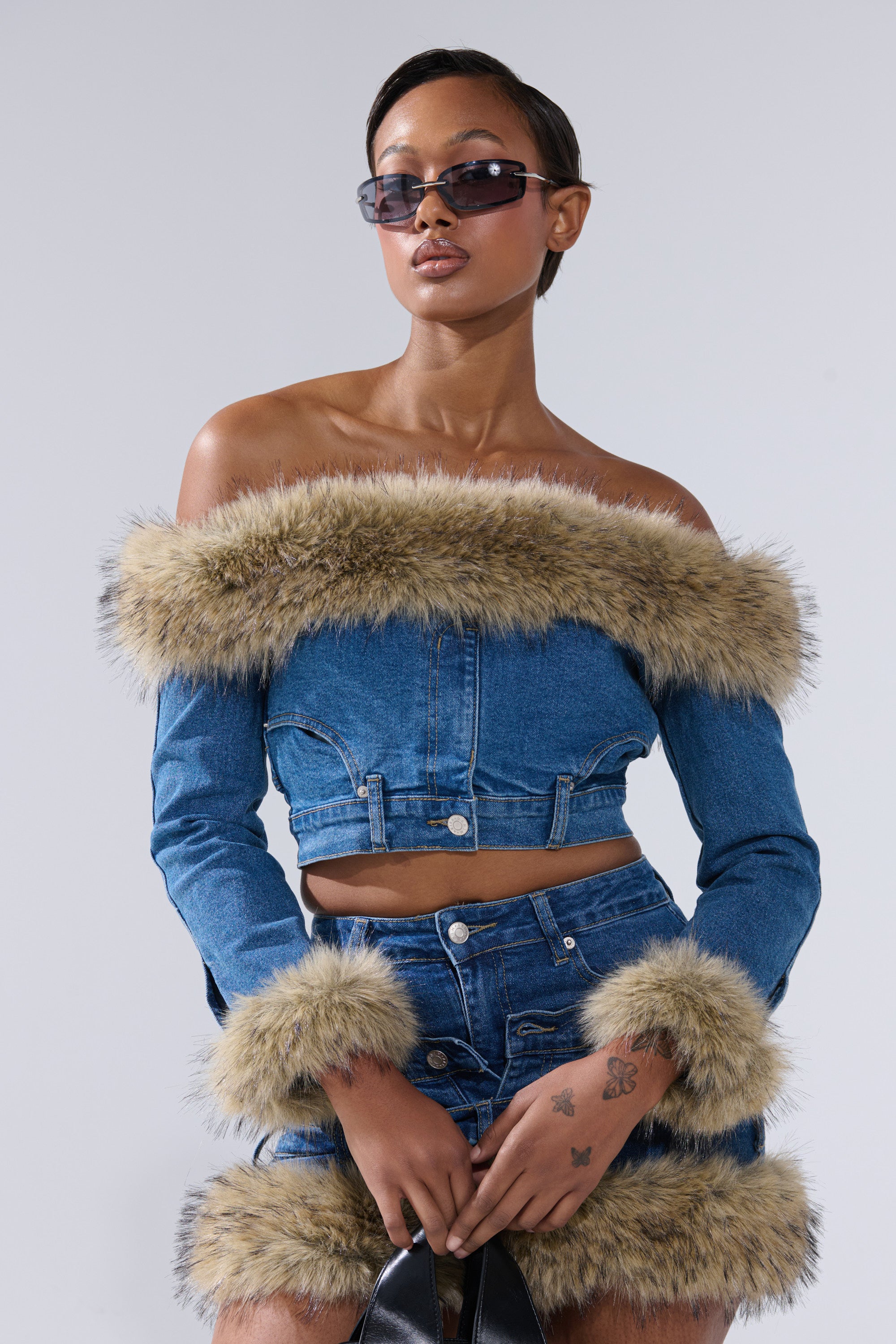 A woman with short hair and sunglasses poses confidently in the FLUFFY OFF THE SHOULDER LONG SLEEVE TOP paired with faux fur trim shorts, holding a black bag against a light gray background.