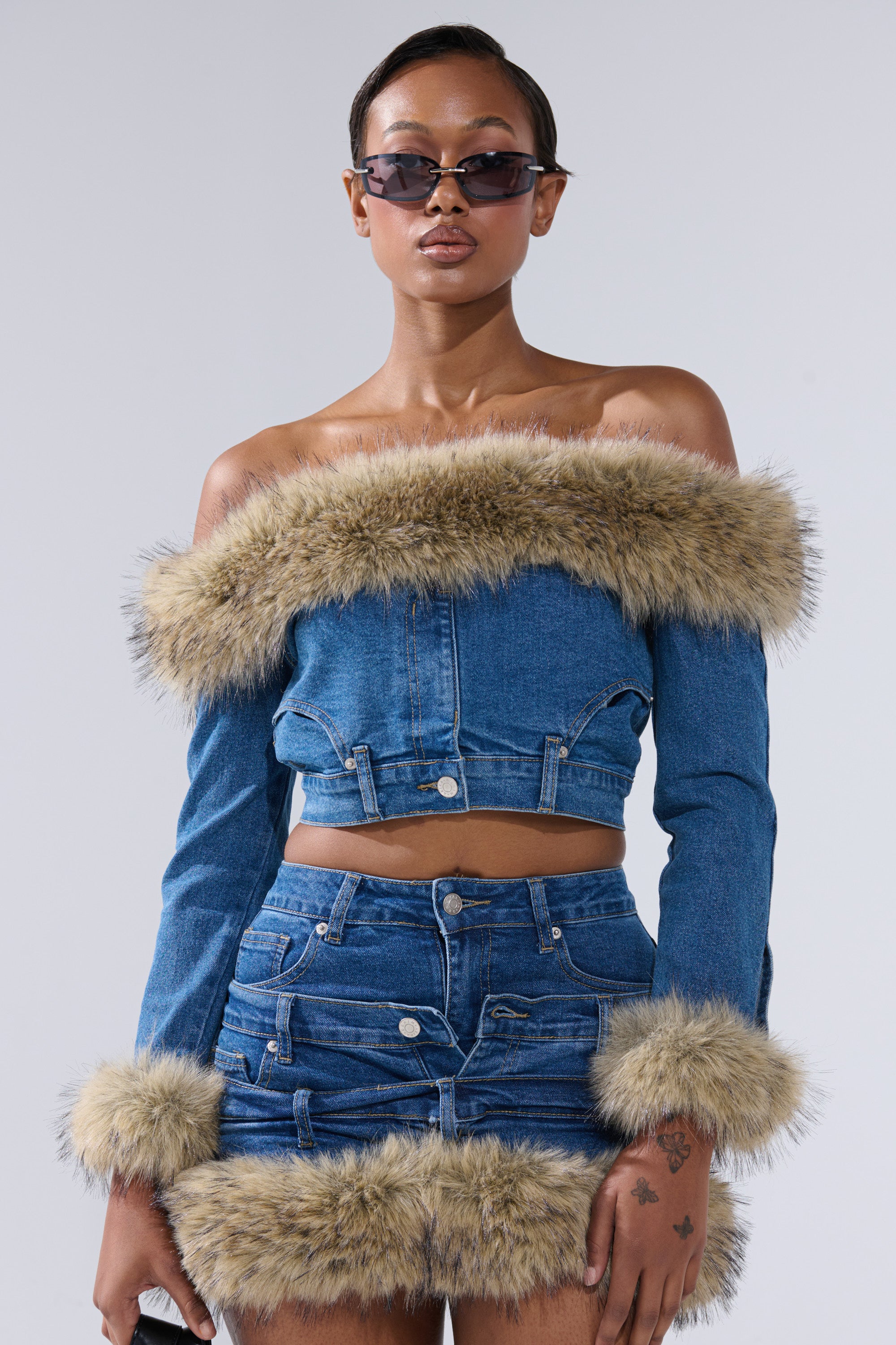 Wearing sunglasses and a FLUFFY OFF THE SHOULDER LONG SLEEVE TOP with tan faux fur trim and a matching mini skirt, a person stands against a plain light background.