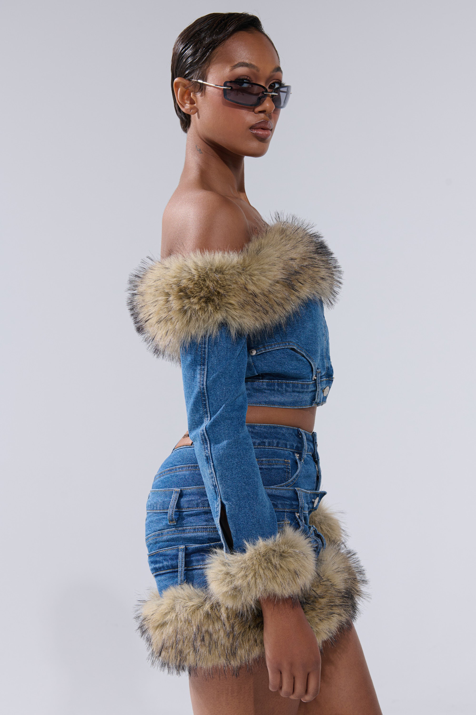 A woman poses confidently in the FLUFFY OFF THE SHOULDER LONG SLEEVE TOP with a matching denim mini skirt, both featuring faux fur trim. She completes her look with trendy sunglasses and glances over her shoulder against a plain background.