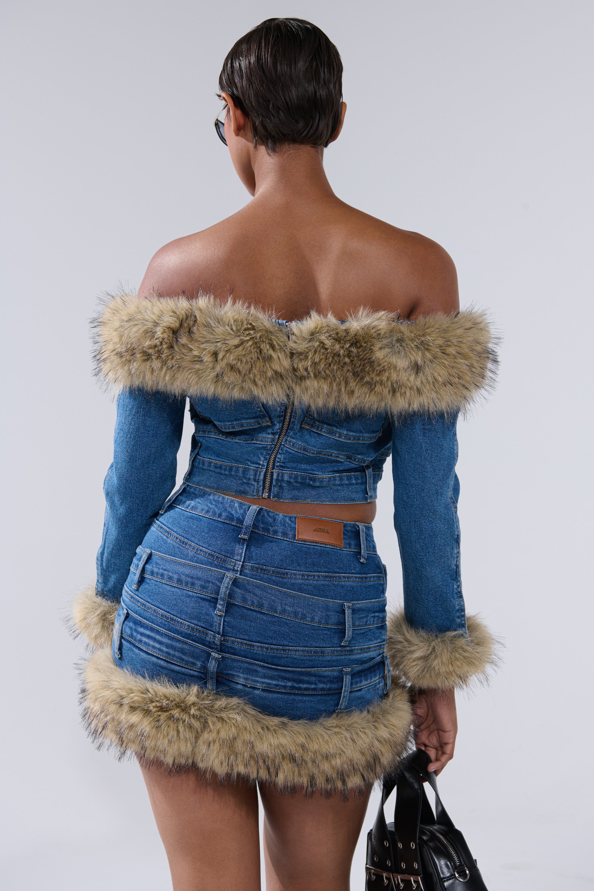 A person with short dark hair models the FLUFFY OFF THE SHOULDER LONG SLEEVE TOP in blue denim, featuring faux fur trim on the neckline, sleeves, and hem. They face away from the camera, holding a black handbag against a plain background.