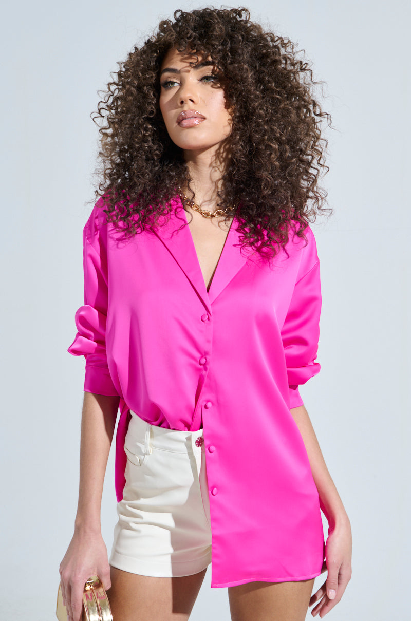 A person with curly hair is wearing a GEO SATIN LONG SLEEVE BLOUSE and white shorts, holding a small gold purse against a plain, light-colored background.