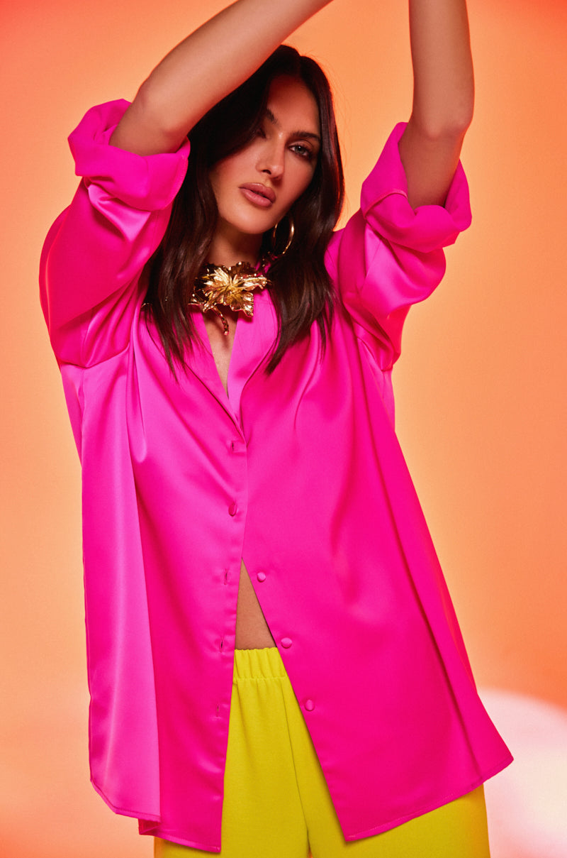 A woman poses with arms raised, wearing the GEO SATIN LONG SLEEVE BLOUSE in bright pink and yellow pants against an orange background. She has loose, wavy hair and accessorizes with a chunky gold necklace.