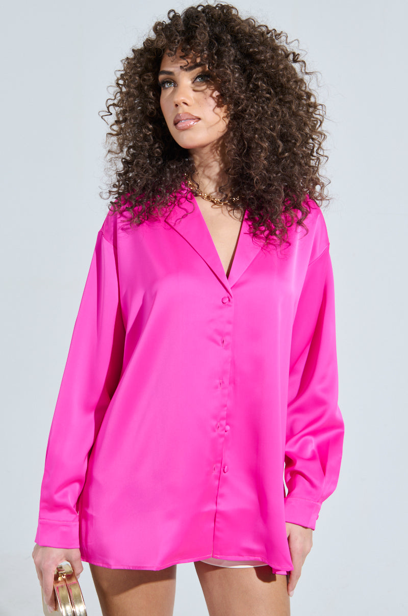 A woman with curly hair wears the vibrant GEO SATIN LONG SLEEVE BLOUSE in pink, glancing upward. She holds a small gold accessory in her left hand against a simple, light gray backdrop.