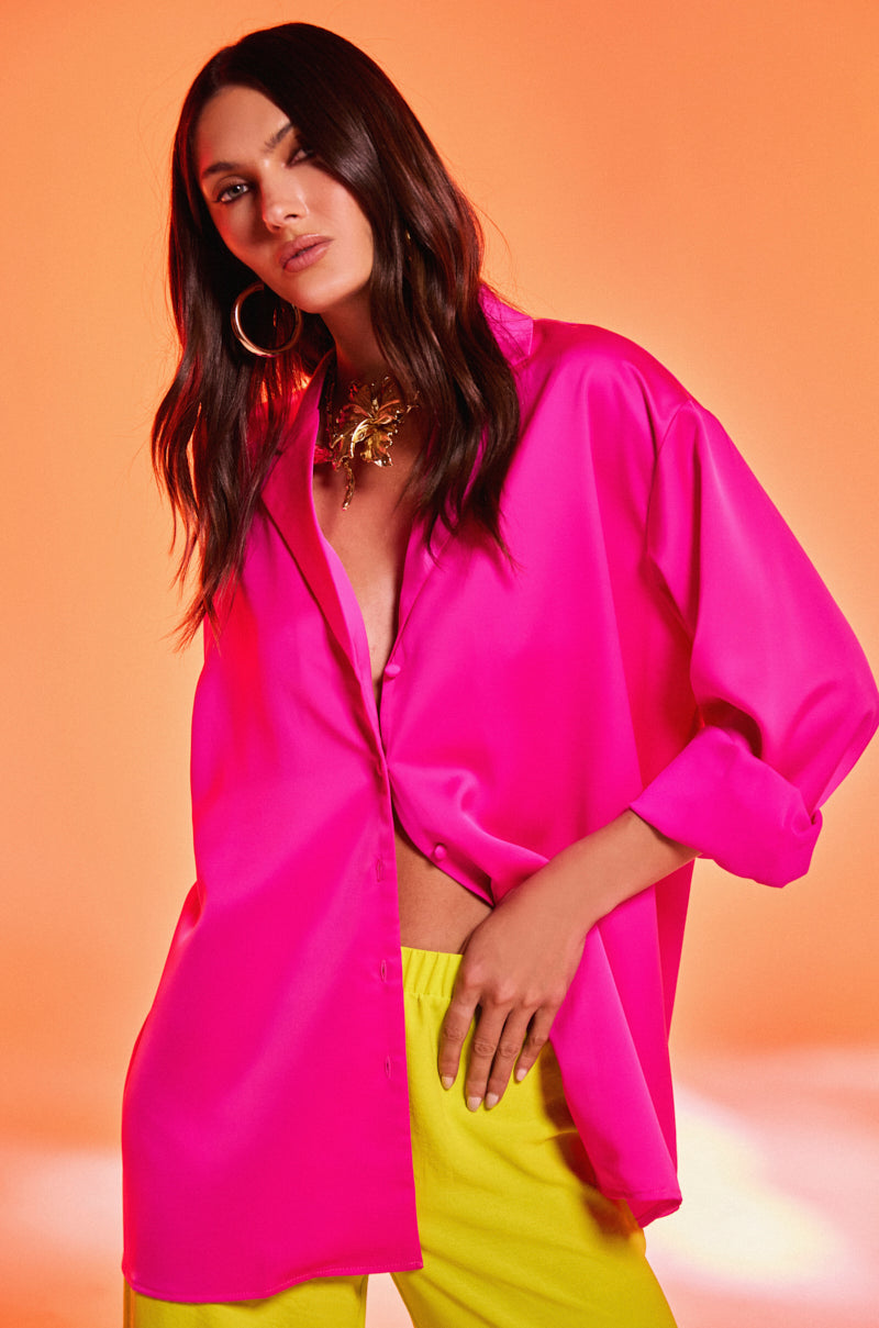 A woman with long dark hair and a confident expression poses against an orange background, wearing the bright pink GEO SATIN LONG SLEEVE BLOUSE, yellow pants, large hoop earrings, and a chunky gold necklace.