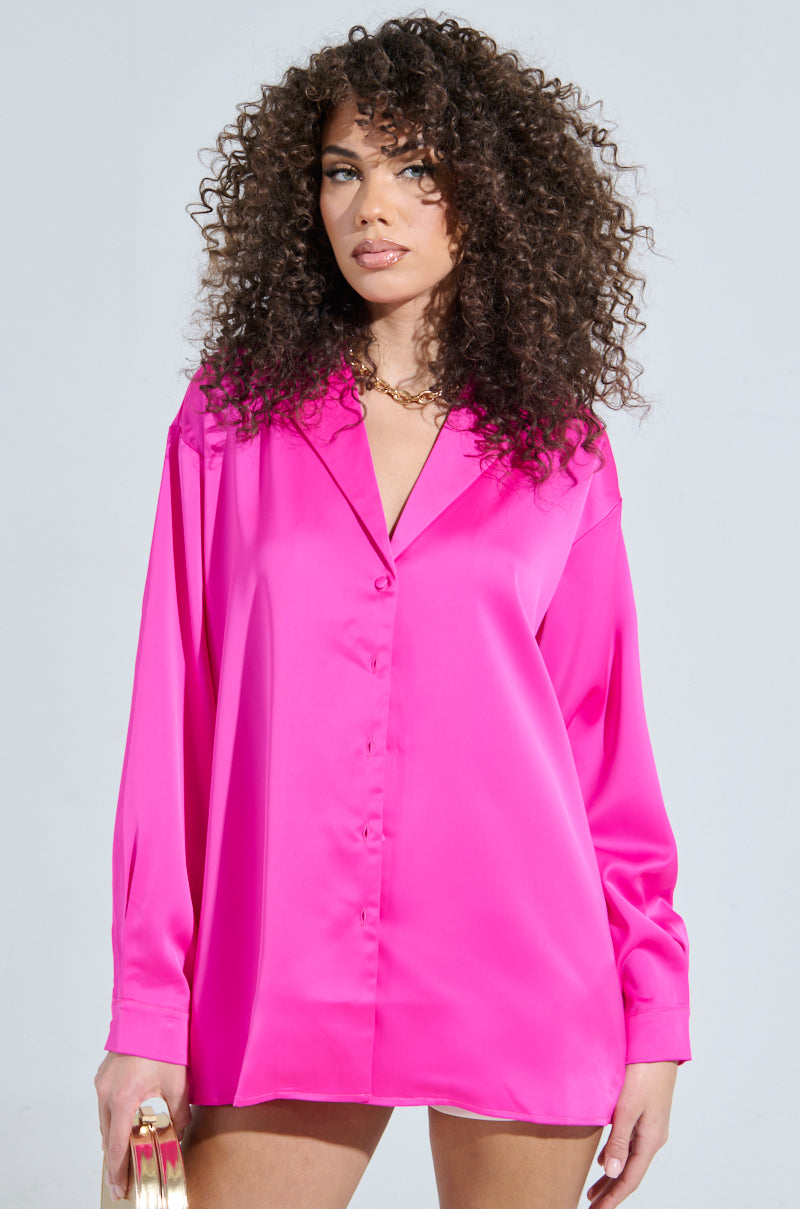 A person with curly hair wears the vibrant GEO SATIN LONG SLEEVE BLOUSE in pink, paired with a small gold handbag. They stand against a light gray background, radiating style and confidence.