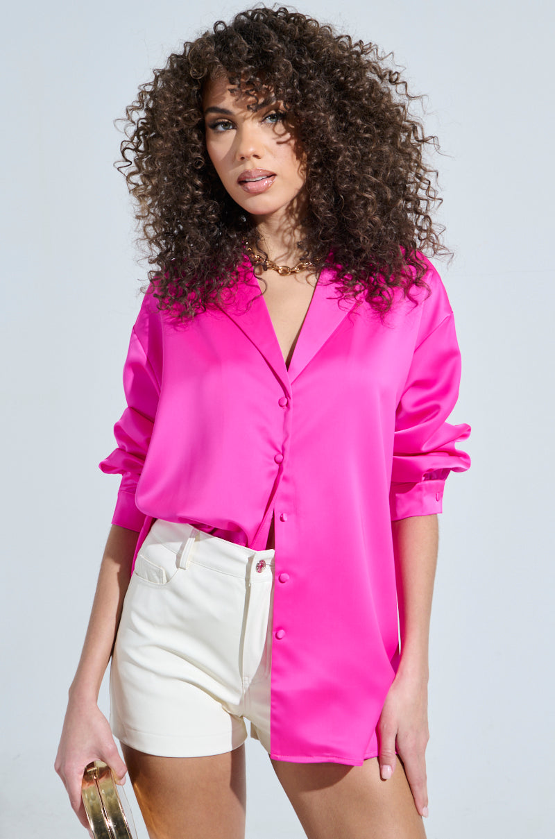 A woman with curly hair wears a GEO SATIN LONG SLEEVE BLOUSE in bright pink and white shorts, standing against a plain background and holding a gold purse.