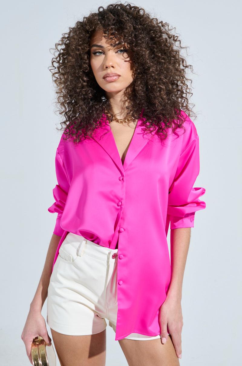 A person with curly hair wears a bright pink Geo Satin Long Sleeve Blouse and white shorts, holding a small gold-toned bag. The plain background accentuates their stylish outfit.