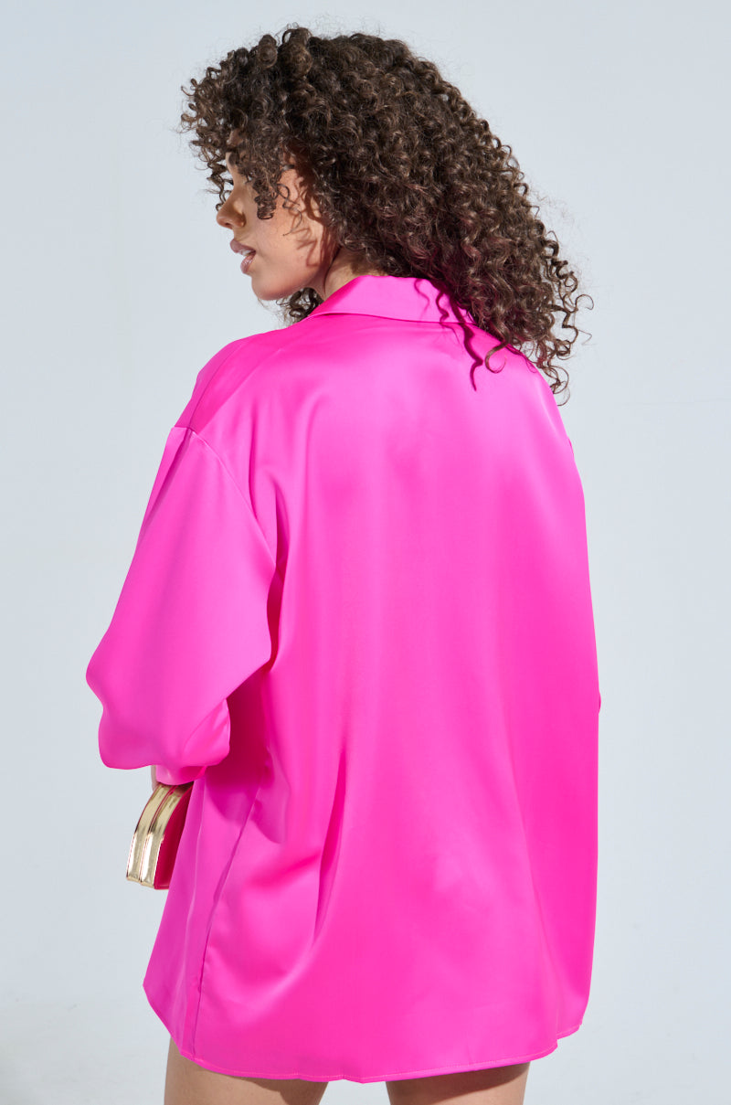 A person with curly hair stands with their back visible, wearing a bright pink oversized GEO SATIN LONG SLEEVE BLOUSE that resembles a Disco Night Top. The background is plain and light-colored, and they hold a partially visible metallic accessory in one hand.