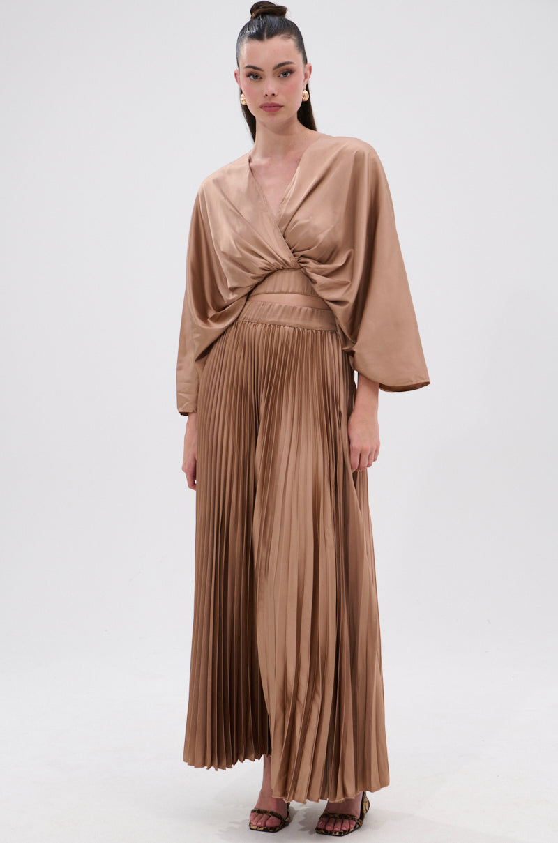 A woman poses before a light backdrop wearing the MILLIE MOCHA MOUSSE SATIN MAXI SKIRT, featuring accordion pleats and a high-rise A-line silhouette, paired with a wrap-style top and her hair in a top knot. 6