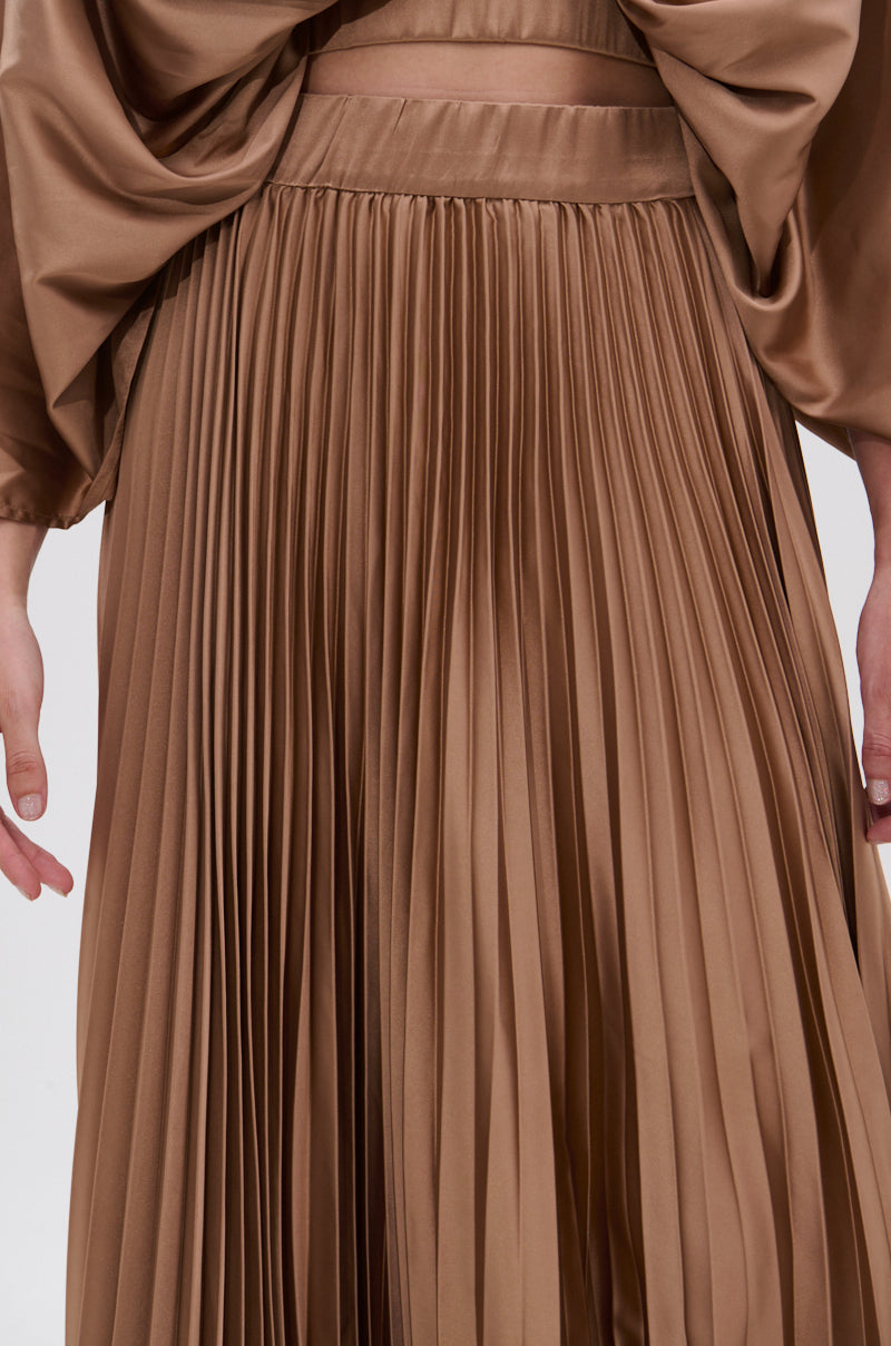 Close-up of a person wearing the MILLIE MOCHA MOUSSE SATIN MAXI SKIRT, showcasing its high-rise A-line silhouette and elegant accordion pleats; their hands rest at their sides against a plain white background. 7