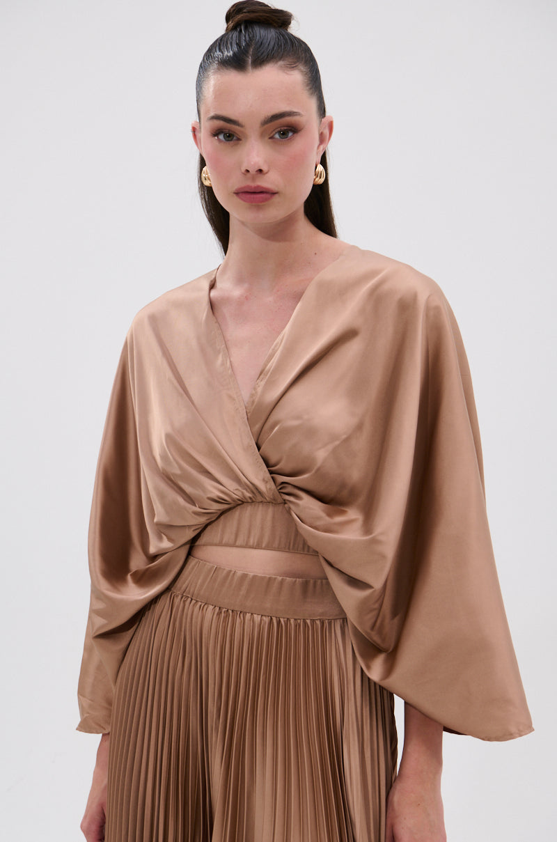 A woman models the MILLIE MOCHA MOUSSE SATIN BLOUSE, featuring draped satin fabric in a shiny mocha hue, paired with a pleated skirt. She poses against a plain light background with her dark hair in a high ponytail and gold hoop earrings. 0