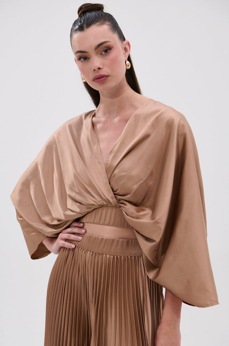 A woman stands against a plain background in the MILLIE MOCHA MOUSSE SATIN BLOUSE, featuring a shiny, light brown pleated design with a deep surplice neckline and wide draped sleeves. She wears her hair in a high ponytail with gold hoop earrings. 1