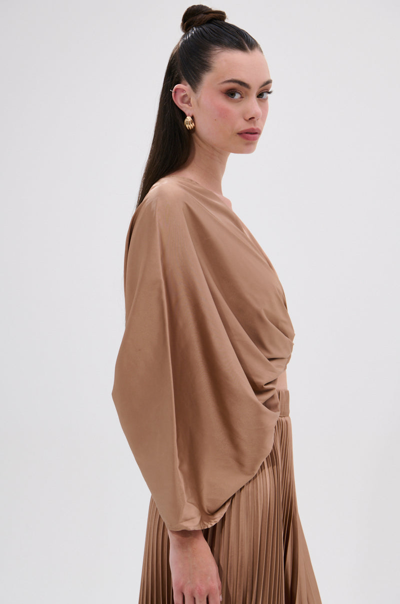 A woman with long dark hair in a topknot, gold hoop earrings, and the MILLIE MOCHA MOUSSE SATIN BLOUSE featuring batwing sleeves and a deep surplice neckline stands against a plain light background, paired with a matching pleated skirt. 2