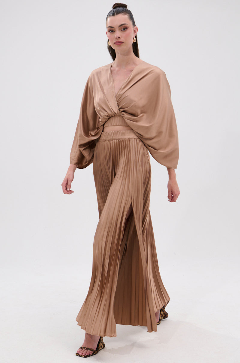A woman walks confidently against a plain white background wearing the MILLIE MOCHA MOUSSE SATIN BLOUSE, featuring a deep surplice neckline paired with matching high-waisted, pleated wide-leg pants. 4