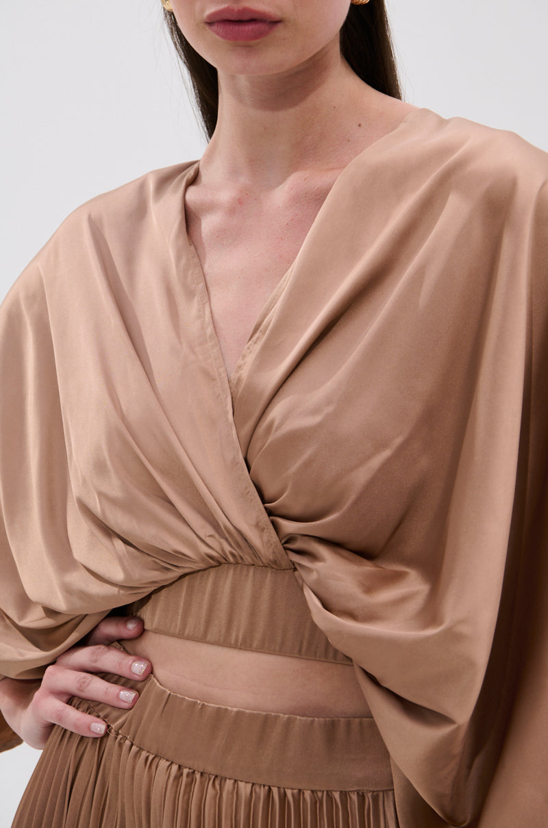A woman models the MILLIE MOCHA MOUSSE SATIN BLOUSE, a silky mocha long-sleeve crop top with a deep V-neck and pleated details. The image is cropped at her nose and midsection. 6