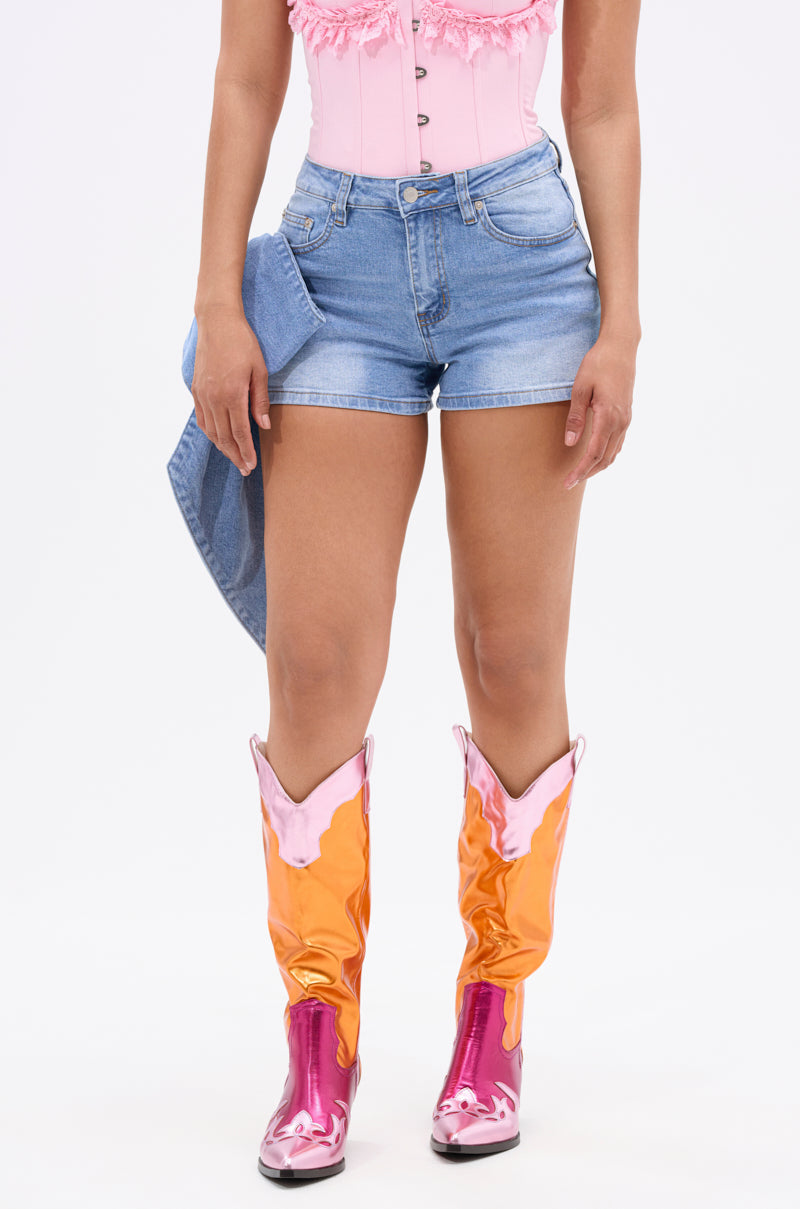 A person stands against a plain white background wearing the YOUR BIGGEST PRESENT BOW DETAIL DENIM SHORT with a pink ruffled top, shiny orange and pink cowboy boots, and holding a denim jacket at their side. 3