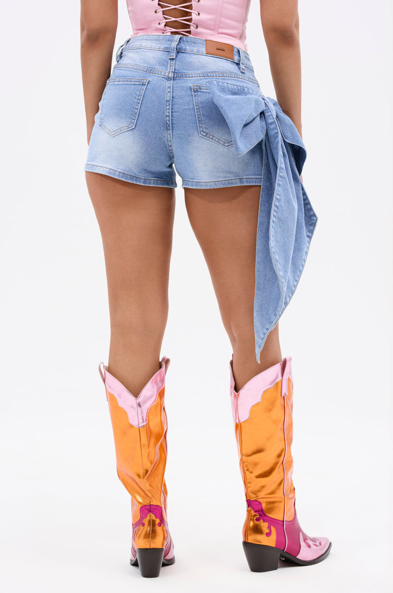 A woman faces away in the YOUR BIGGEST PRESENT BOW DETAIL DENIM SHORT, styled with a pink lace-up top and bright orange-pink cowboy boots against a plain white background. 5