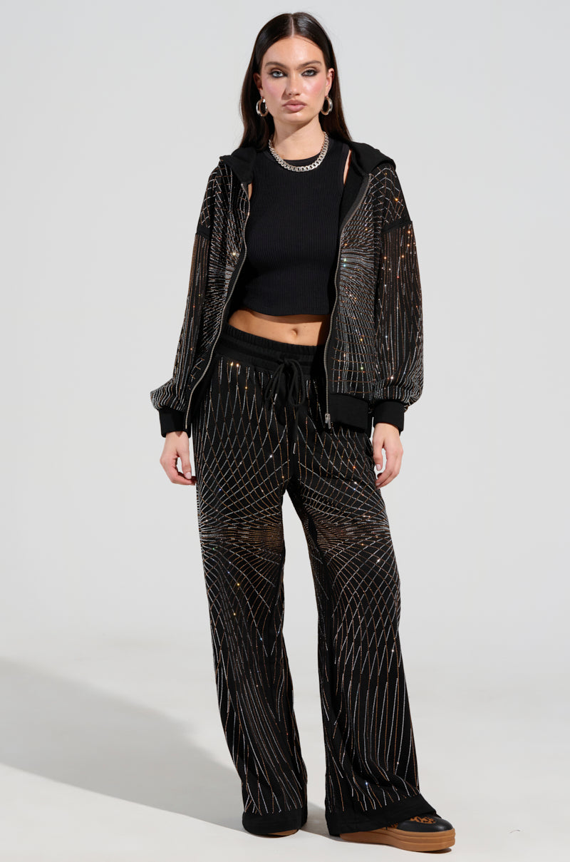 A woman poses in the LIGHTS, CAMERA, ACTION! EMBELLISHED WIDE LEG SWEAT PANT with rhinestone detailing, accompanied by a matching black patterned jacket and crop top. She pairs her outfit with black shoes, letting her long, straight hair fall naturally as she stands against a simple gray background.