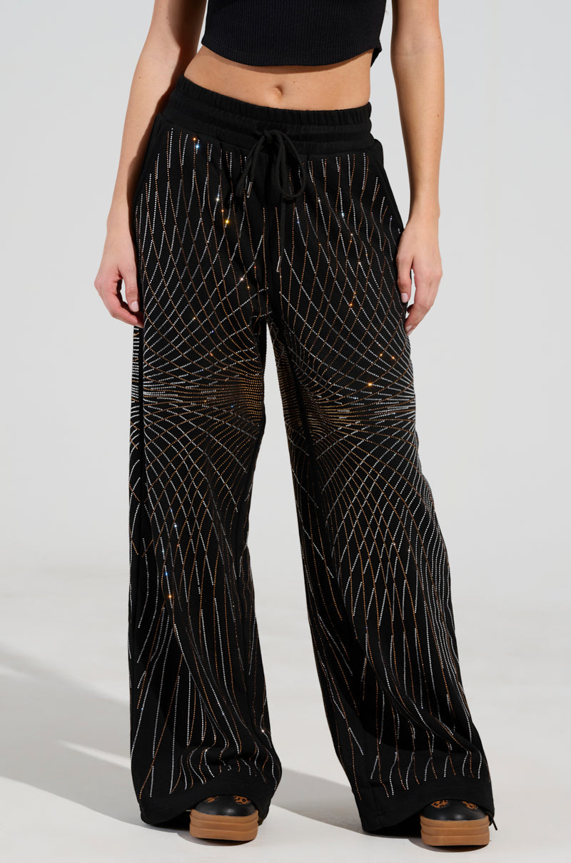 A person wearing LIGHTS, CAMERA, ACTION! EMBELLISHED WIDE LEG SWEAT PANTS with a geometric gold and blue pattern and a high-rise waistband stands against a light background. They pair these pants with a black cropped top and brown shoes, with their hands resting casually by their sides.